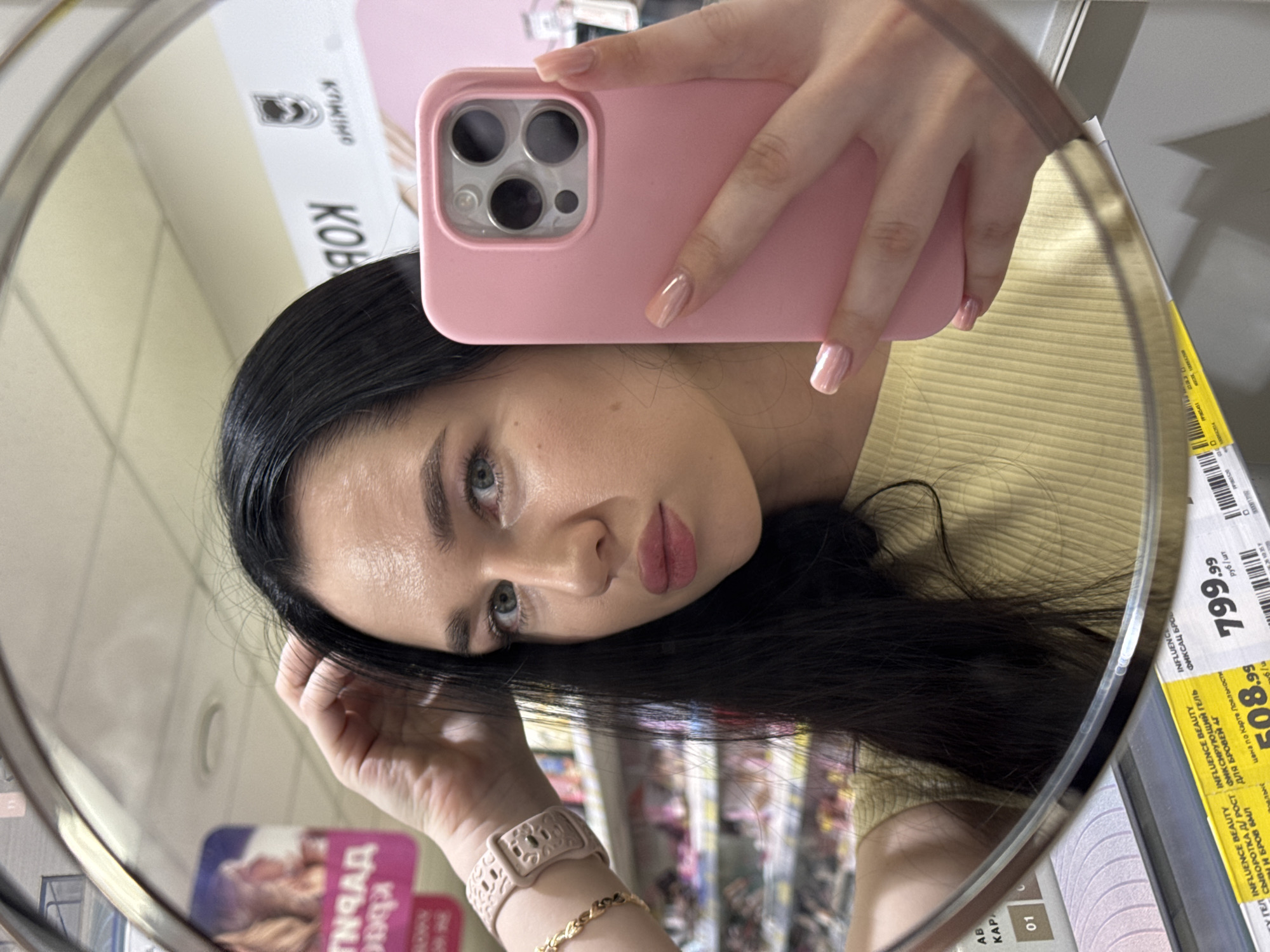 A young woman with long, dark hair and light blue eyes is taking a mirror selfie in a brightly lit store. She is wearing a yellow ribbed t-shirt and has a pink phone case in her right hand. She is also wearing a light pink watch on her left wrist and a gold bracelet. The background of the mirror reflection shows shelves stocked with various products, likely in a cosmetics or beauty store. Some price tags and product packaging are visible, including a yellow tag with Russian text. The overall environment appears to be indoors, possibly a supermarket or a dedicated beauty retailer, common in urban areas like Novosibirsk, Russia.