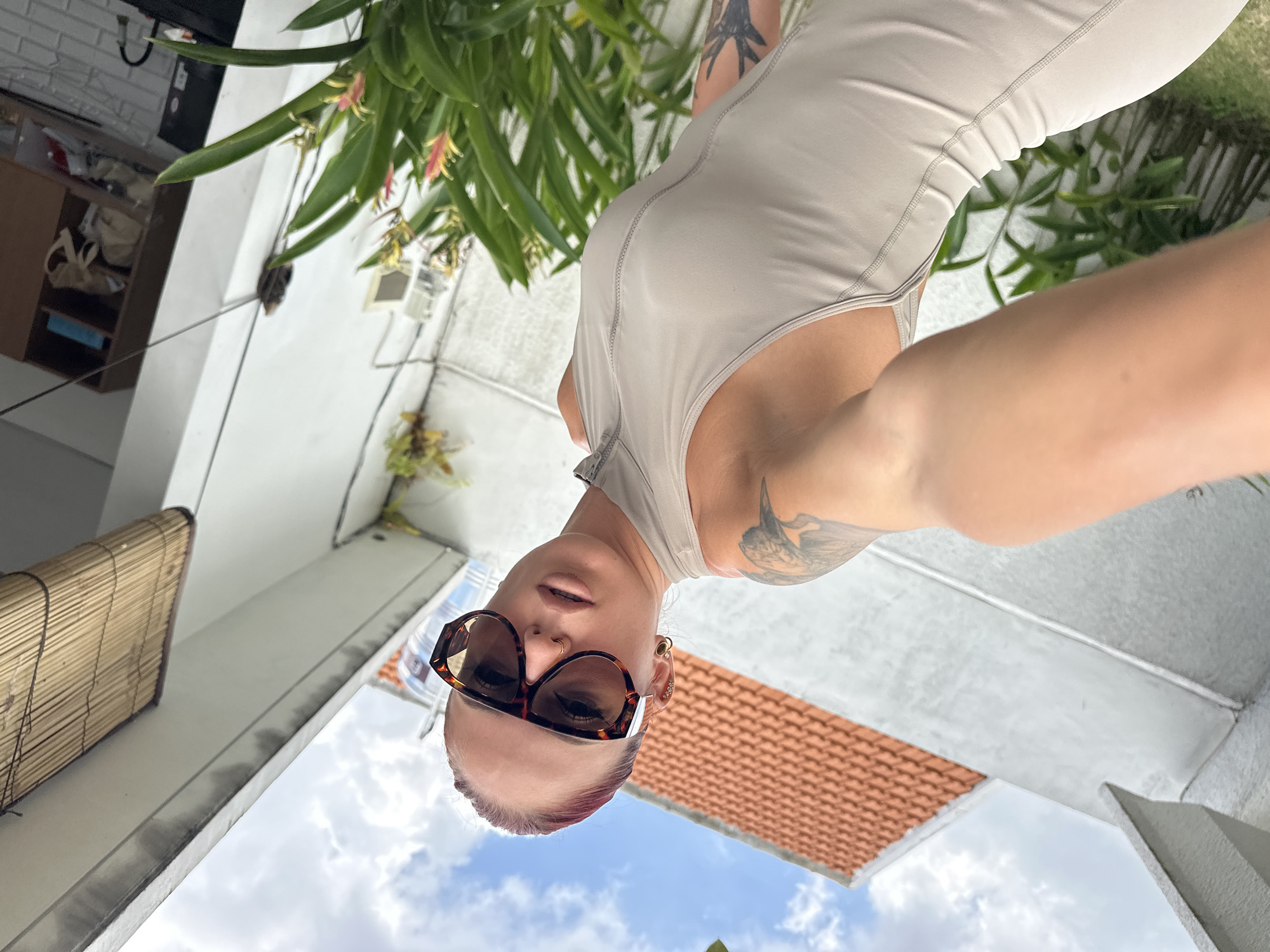The image depicts a woman taking a selfie in what appears to be a residential area. The environment includes greenery, a building with a red-tiled roof, and a cloudy sky. The location is potentially in North Kuta, Indonesia, based on the architectural style.