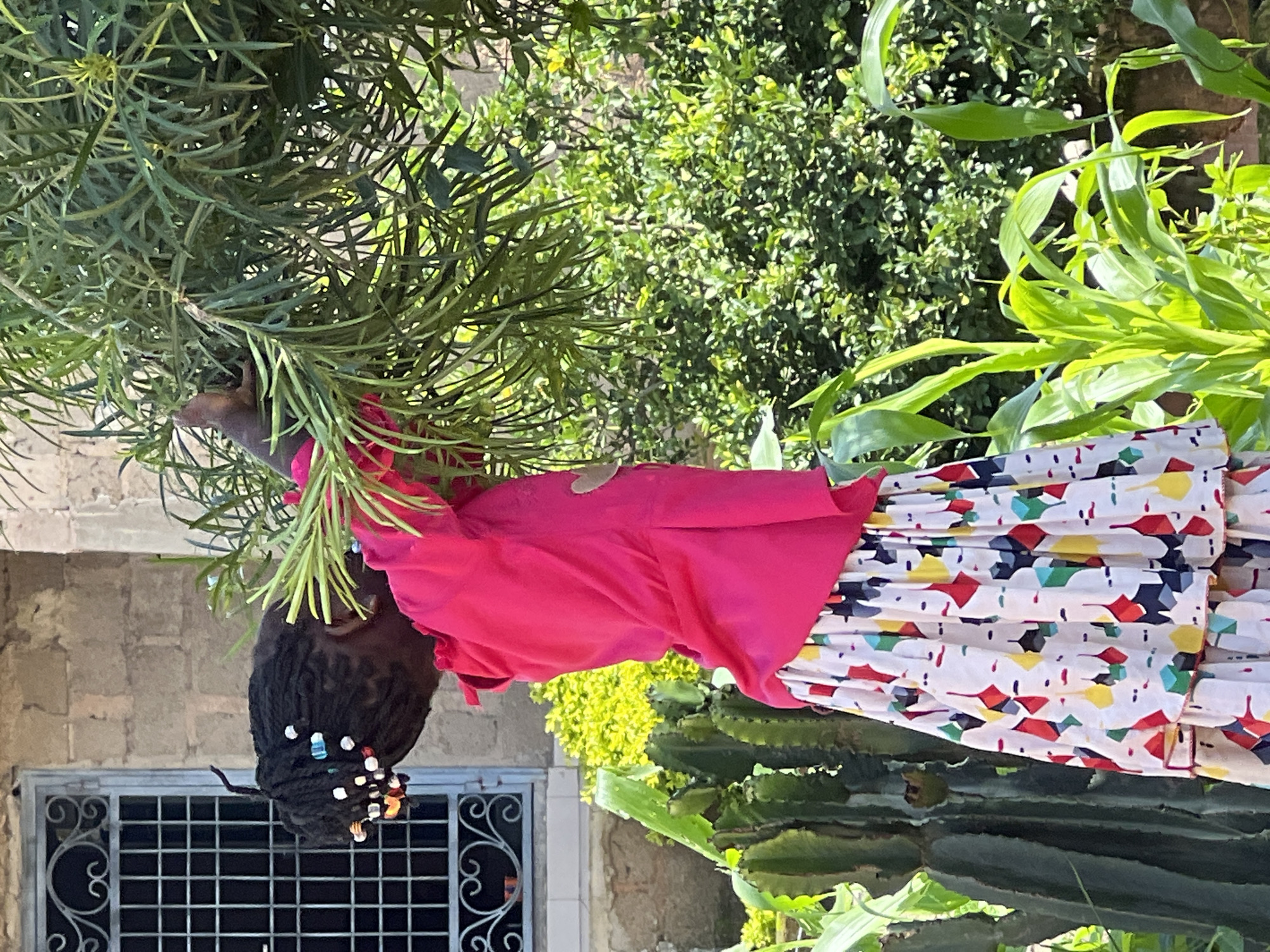 A young Black girl with braided hair adorned with beads is reaching into a leafy green tree. She is wearing a bright pink long-sleeved shirt and a white tiered skirt with a colorful geometric pattern. Behind her, a stone building with a barred window is visible, along with lush greenery, including cacti and corn stalks. The scene appears to be outdoors, likely in a garden or yard, bathed in sunlight. This image was taken in the city of Jos, Nigeria.
