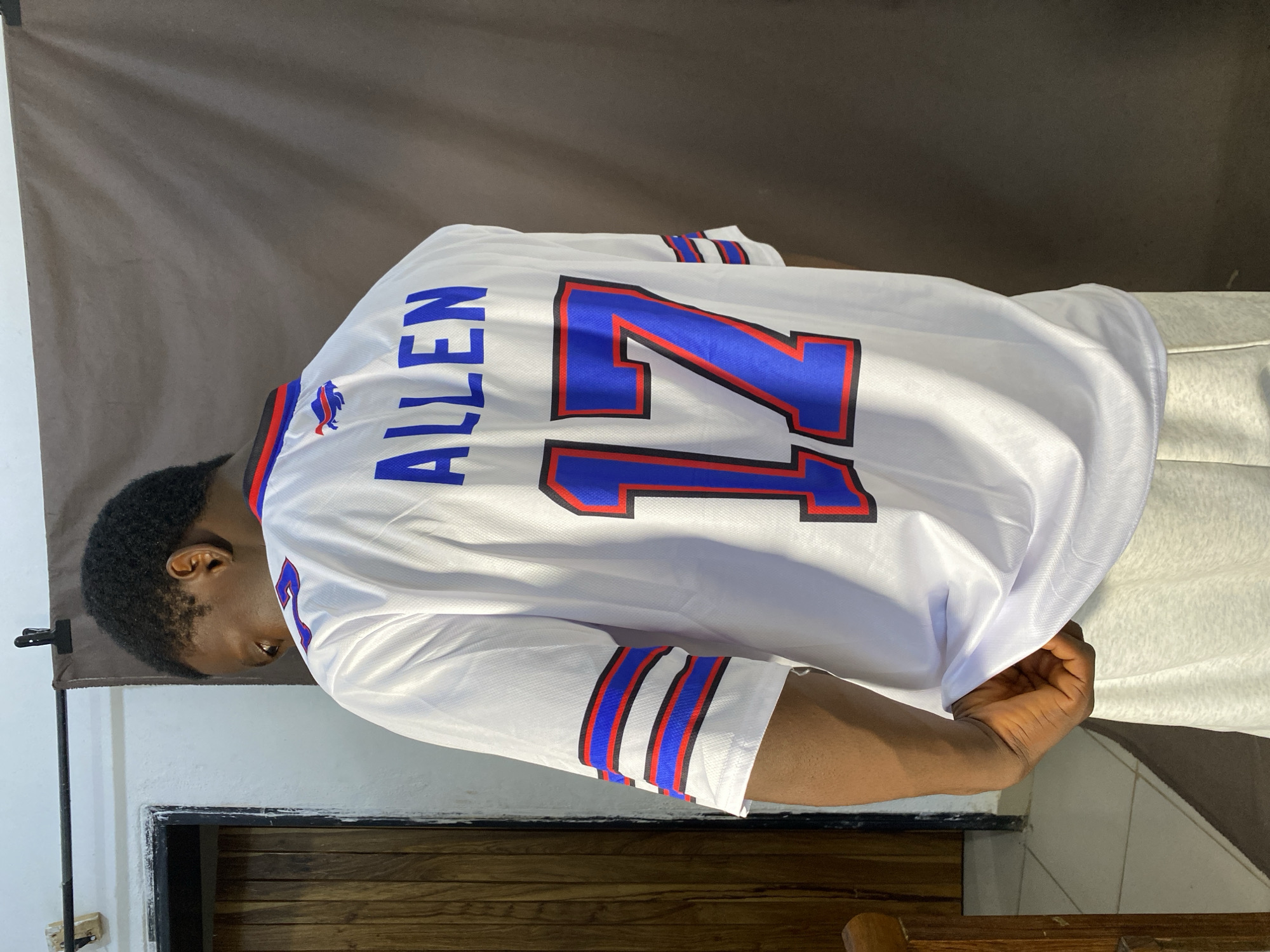 A young Black male stands with his back to the camera, wearing a white Buffalo Bills jersey with "ALLEN" and the number "17" in blue with red and black trim. The jersey has blue and red stripes on the sleeves. He is wearing light gray sweatpants. He is standing in front of a dark brown backdrop. To his left is a wooden door and a white wall. The floor is tiled. The scene appears to be in a studio or indoor setting in Bukuru, Nigeria.