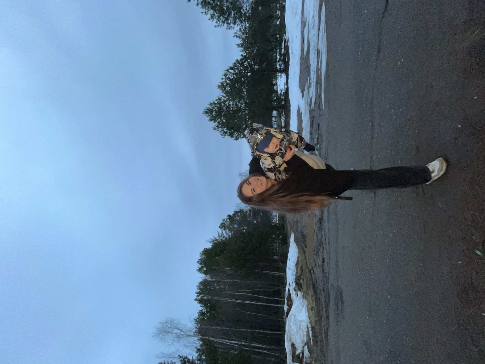 The image shows a woman holding a young child in a wooded area in Chaikovsky, Russia. The woman is wearing black pants and jacket with light colored shoes. The child is wearing a patterned jacket and hat. There is snow on the ground. The sky is overcast. The woman and child are standing on what appears to be a road or paved area. Trees line the background.