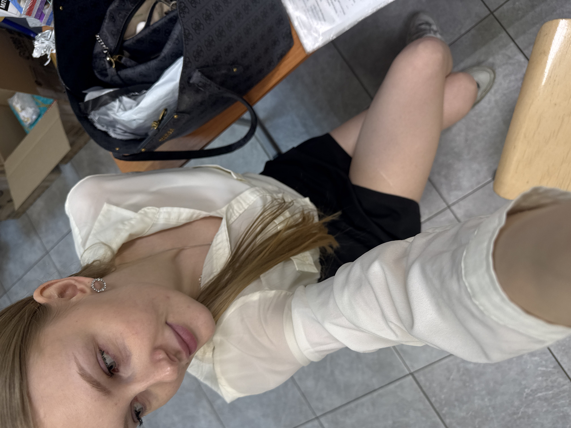 A young woman with fair skin and light brown hair is taking a selfie. She is wearing a white blouse and a black skirt. The background includes a black Guess handbag on a wooden surface, a cardboard box, a wooden chair leg, and a tiled floor. The location is likely indoors in Perm, Russia.