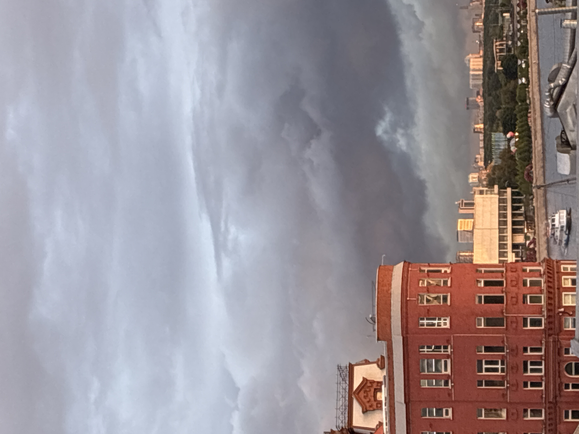 The image depicts a cloudy sky over Moscow, Russia, with buildings in the foreground. The sky transitions from light gray clouds to darker, potentially stormy clouds. A red brick building with numerous windows is prominently featured, its architecture suggesting a historical or residential structure. In the distance, other buildings and trees contribute to the urban skyline.