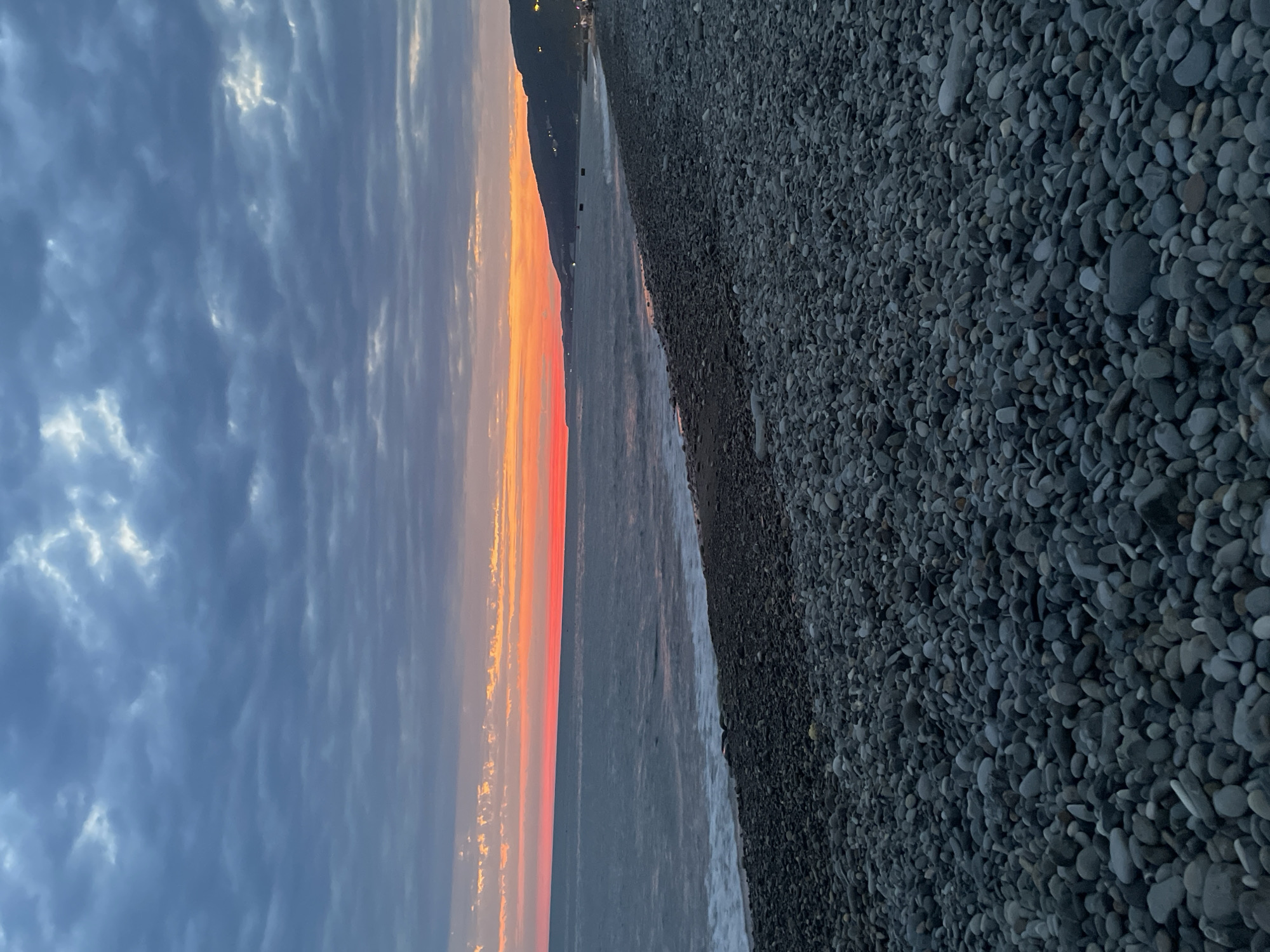 The image depicts a pebbled beach at dusk. The sky is filled with dramatic, gray clouds, with a sliver of vibrant orange and pink light on the horizon where the sun has set. The sea is a deep blue-gray, with gentle waves lapping at the shore. The beach is densely covered with smooth, dark gray and black pebbles of various sizes. In the distance, along the coastline, a silhouette of hills is visible, dotted with small lights that suggest a settlement. The overall atmosphere is serene and tranquil, characteristic of twilight by the sea.