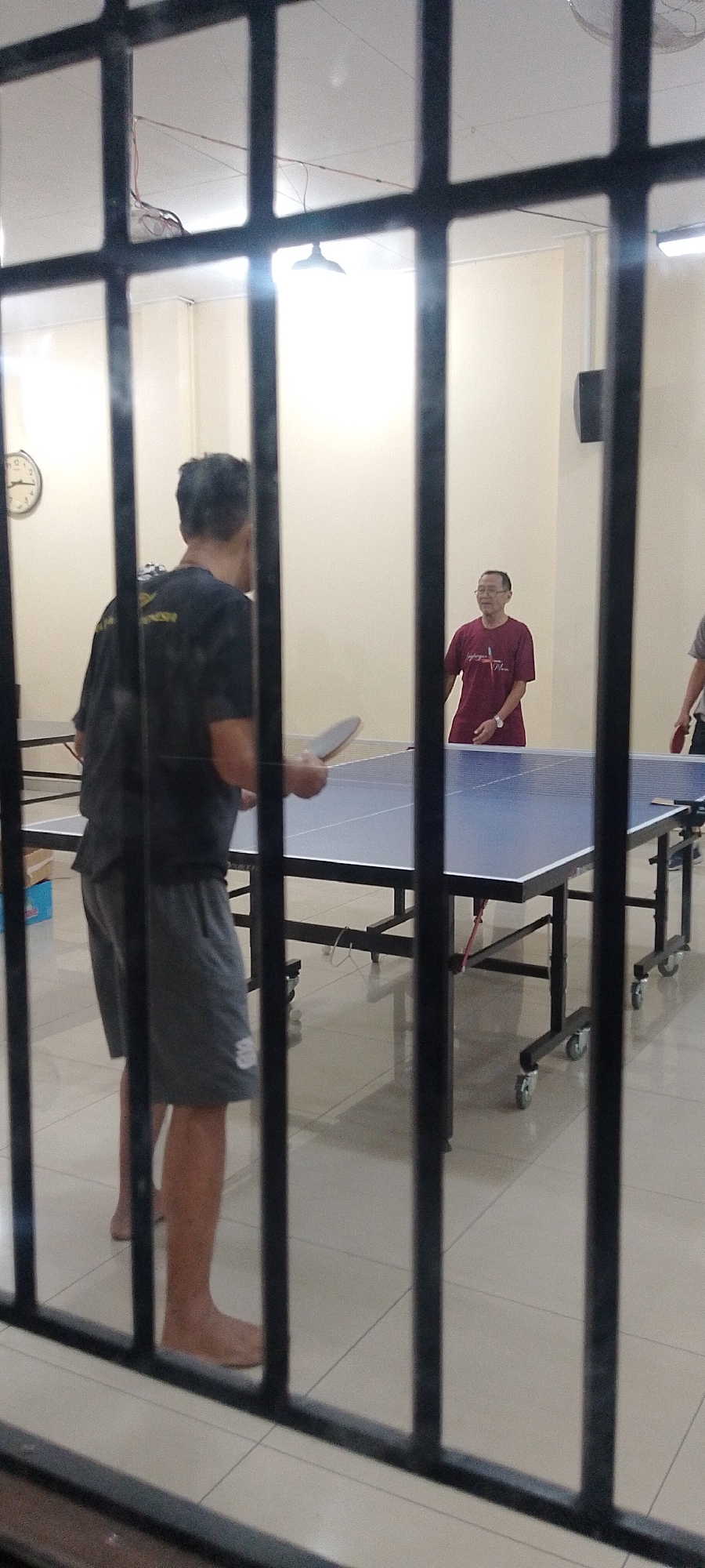 Two individuals are engaged in a table tennis match, partially obscured by a dark metal grate in the foreground, within an indoor facility. The person on the left, wearing a black t-shirt and gray shorts, is facing away from the camera, holding a table tennis paddle. The person on the right, wearing a maroon t-shirt, is facing the camera and also holding a paddle. A blue table tennis table is positioned between them. The room has beige walls and a tiled floor. A circular clock is visible on the left wall. The image appears to be taken in Kudus, Indonesia, based on the context provided.