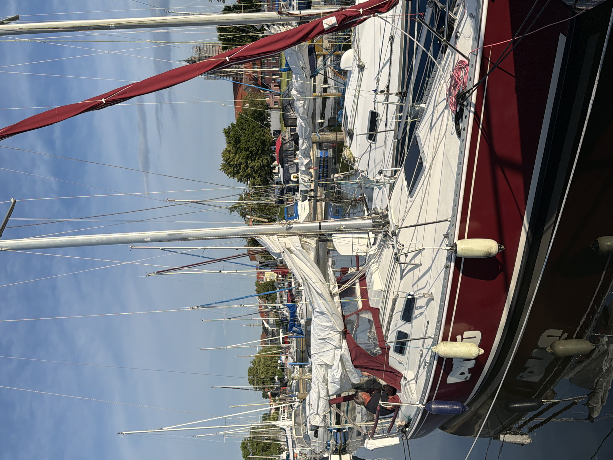 This image captures a marina in Ribnitz-Damgarten, Germany, on a clear day with a partly cloudy sky. The foreground is dominated by a red and white sailboat with its sail furled in a burgundy cover. A man, appearing to be in his 60s with gray hair, is seated on the deck of the boat, facing away from the camera and looking towards the interior. The boat is equipped with several white fenders hanging from its sides, and its reflection is visible in the calm water. In the background, numerous other sailboats are moored, their masts creating a dense forest against the sky. Beyond the marina, buildings with red roofs and green trees are visible, suggesting a town or residential area adjacent to the waterfront. The overall impression is one of a peaceful, organized harbor.