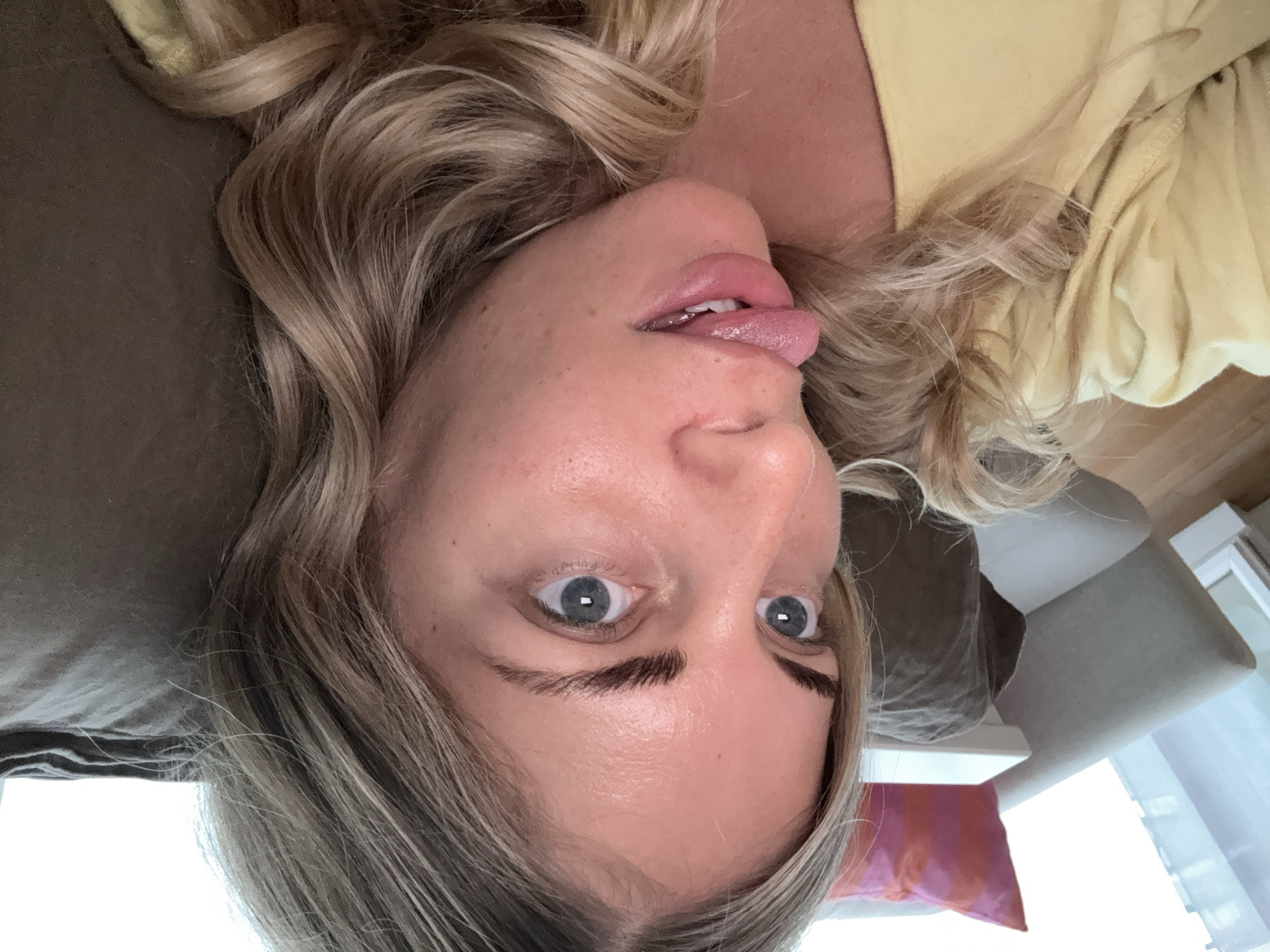 A woman with blonde hair is lying down, possibly on a couch, sticking her tongue out. She has fair skin and blue eyes. Her clothing appears to be a light yellow color. The background includes furniture like a couch and cushions. There is no information in the image to determine the location as Paris, France.