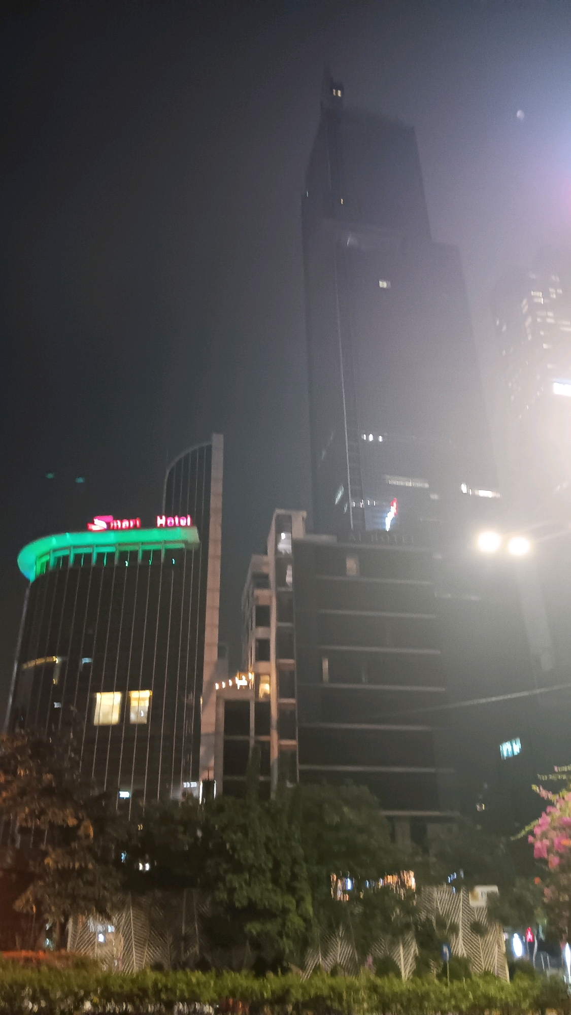 This is a vertical, night-time photograph of a city skyline in Central Jakarta, Indonesia. The most prominent structure is a tall, dark skyscraper that dominates the right side of the frame, its upper sections obscured by haze and bright lights. To the left of this skyscraper, there is a curved building with a green, illuminated top that spells out "Smart Hotel" in red neon letters. In front of and between these buildings, there are several lower structures, some appearing to be under construction or partially built, with visible windows and lighting. Trees and dense foliage are visible in the foreground, partially obscuring a white, decorative fence-like structure. The overall atmosphere is hazy, with bright lights from the buildings and streetlights reflecting and diffusing in the night air. There are no clearly discernible people in the image.