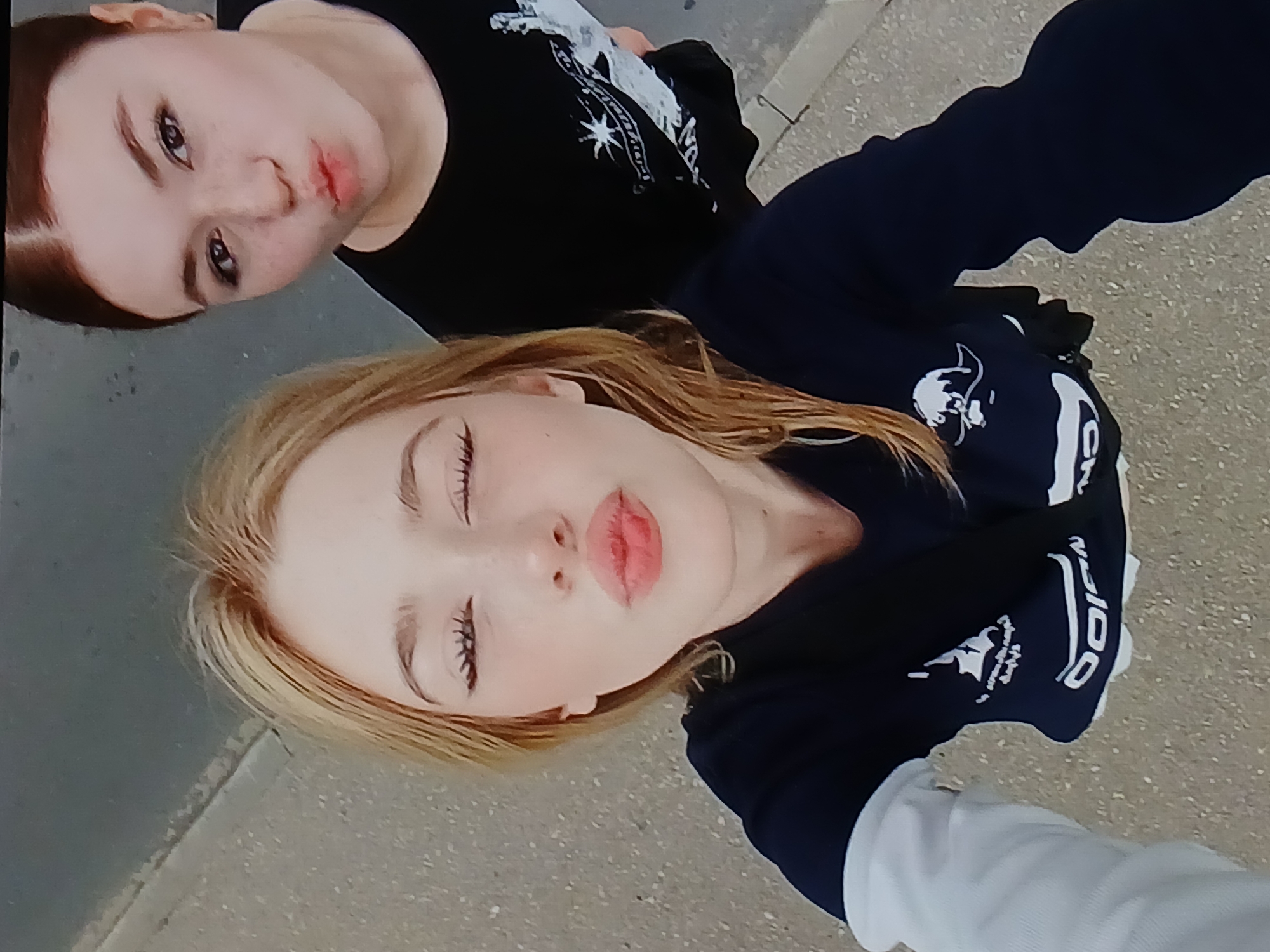 In an outdoor, eye-level, medium shot taken from above, two young women pose for a selfie on a paved surface, possibly a sidewalk, with a grey, speckled texture. The woman in the foreground, with blonde hair and closed eyes, puckers her lips for a kiss. She wears a dark blue long-sleeved shirt with white accents on the sleeves and a graphic print on the chest. The woman behind her, with dark brown hair pulled back and a slight smirk, also pouts her lips. She is wearing a black t-shirt with a white graphic print. The background is blurred, showing a grey surface that transitions to a darker, greyish-blue area at the top, suggesting an urban environment in Dnipro, Ukraine.