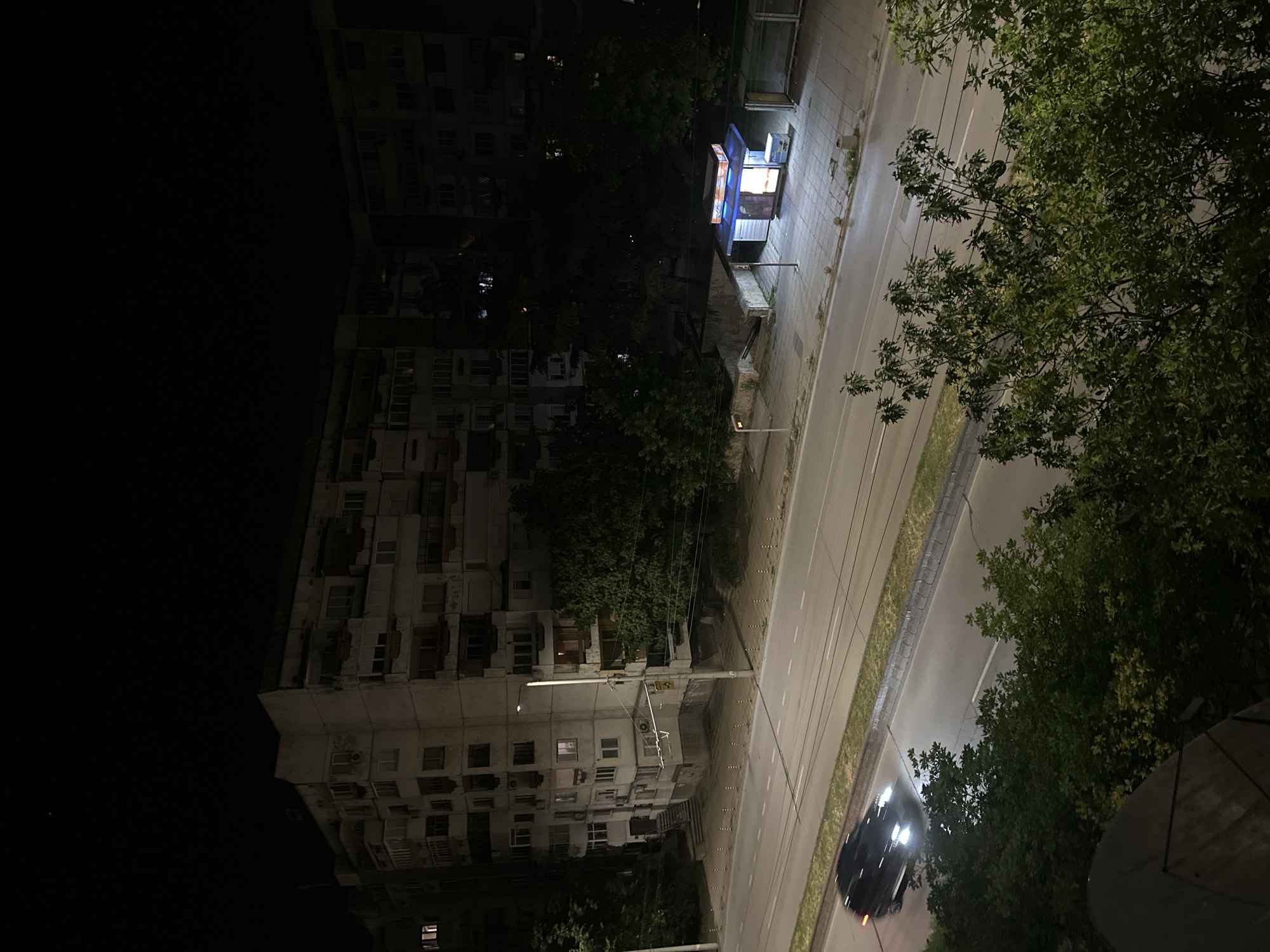 This high-angle nighttime photograph depicts an urban scene within the city of Unknown, Unknown. The primary focus is a street flanked by multi-story residential buildings and various urban infrastructure.
In the background, two large, dilapidated residential buildings with numerous windows and balconies dominate the upper half of the image. These structures appear to be made of concrete or stucco, exhibiting signs of wear and tear. Some windows on the buildings are illuminated, indicating occupancy. Trees are visible in front of and between the buildings, partially obscuring the lower floors. The sky above is completely dark, suggesting a clear night with minimal ambient light beyond artificial sources. A faint light source or celestial body is visible in the upper left of the sky.
A paved, multi-lane road runs horizontally across the mid-ground, featuring white lane markings. On the right side of the street, there is a brightly lit kiosk with a blue roof and prominent signage, possibly indicating a convenience store or newsstand. Adjacent to the kiosk is a bus stop shelter constructed of glass and metal. Further along the sidewalk on the right, there are small concrete posts lining the curb. On the left side of the street, a utility pole with a streetlight stands near one of the residential buildings, partially draped with wires. The sidewalk directly in front of the building on the left features a set of exterior stairs leading to an entrance.
In the foreground, a dark-colored car with illuminated headlights is in motion on the left side of the street, appearing slightly blurred due to movement. Dense green foliage from trees and bushes occupies the lower portion of the image, partially obscuring the view of the street and foreground elements. A dark, curved object, possibly part of a satellite dish, is visible in the bottom left corner. The scene is illuminated primarily by artificial lights from the streetlights, buildings, and the kiosk, casting shadows and highlighting specific areas. No individuals are discernible in the photograph.