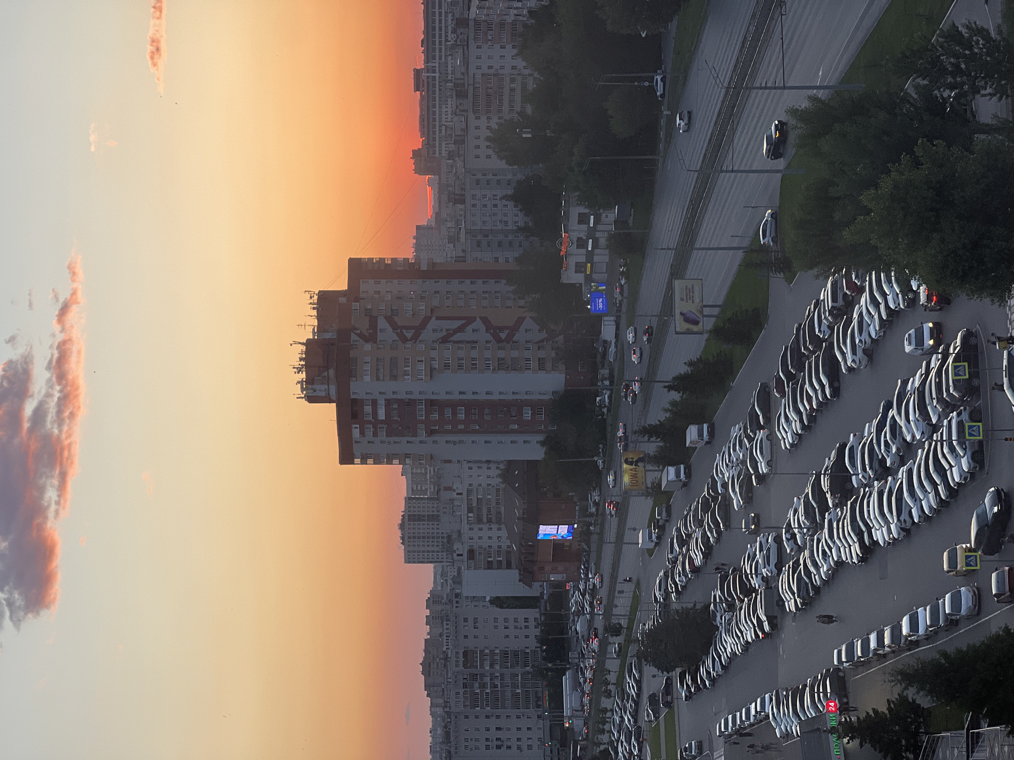 This is a high-angle, eye-level, medium shot of a cityscape during a sunset. The sky is a gradient of orange and pink, with scattered clouds. In the foreground, a large parking lot is filled with numerous cars, mostly white and silver. Adjacent to the parking lot is a multi-lane road with a few cars driving on it. Beyond the road, there is a dense cluster of residential buildings and a tall, reddish-orange apartment building with distinctive architectural features. A large digital billboard is visible on the side of one of the buildings. Trees and greenery are interspersed throughout the scene. The overall impression is of an urban environment at dusk. The scene is likely in Kazan, Russia, given the architectural style and the billboard's text, which appears to be in Cyrillic.
