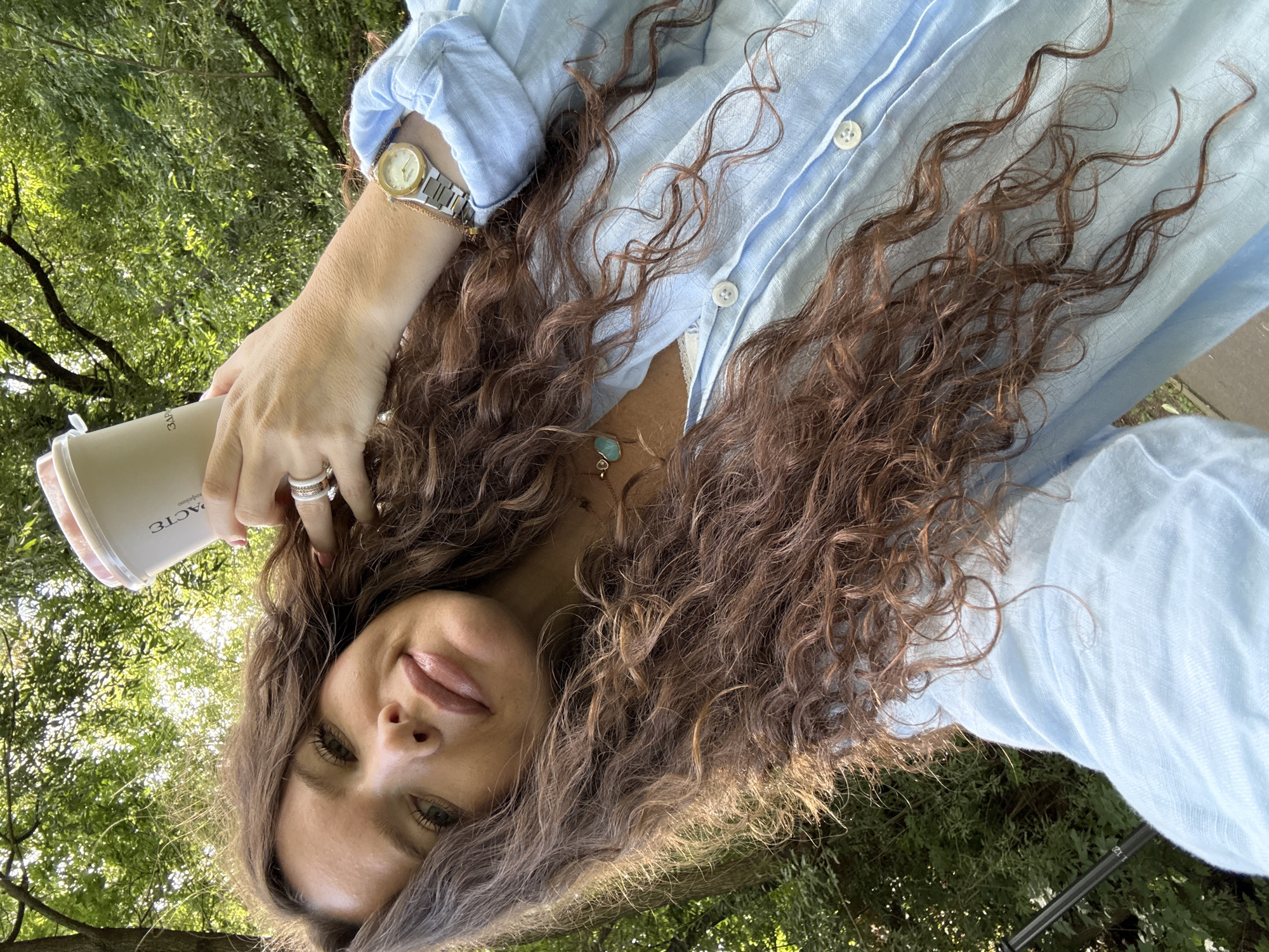 A woman with long, curly brown hair is taking a selfie outdoors, holding a coffee cup in her right hand. She is wearing a light blue collared shirt and a silver watch on her left wrist. She also has rings on her fingers. The background appears to be a park or wooded area with trees and foliage. The lighting suggests it is daytime. There are no visible landmarks that would indicate a specific location within Moscow, Russia.
