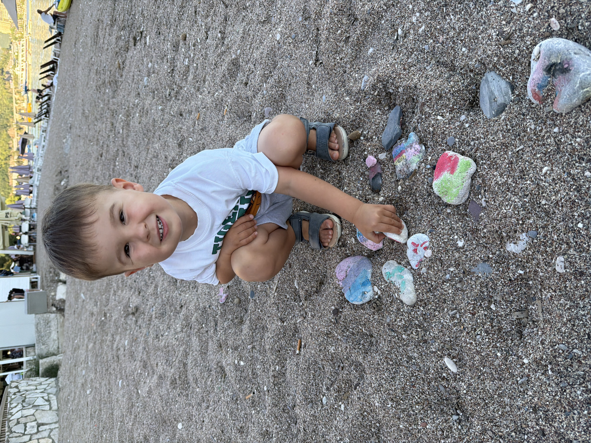 A young Caucasian male child, estimated to be between two and four years old, is depicted crouching on a pebbled beach. He is wearing a white short-sleeved t-shirt with a green graphic, light gray shorts, and gray sandals. He is smiling directly at the viewer, with his right hand reaching towards a cluster of approximately a dozen smooth rocks, many of which are painted with various bright colors including blue, pink, green, red, and white. A discarded cigarette butt is visible on the pebbles to the left of the child. The immediate ground consists of small, mixed-color pebbles and coarse sand. In the background to the left, a stone-clad building, possibly a beach bar or restaurant, is visible with obscured figures. To the right, the beach extends towards a body of water, identified as the Adriatic Sea in Budva, Montenegro. Several sun loungers and closed beach umbrellas are arranged along the shoreline, and multiple boats are anchored in the calm water further out. Additional human figures are present in the distant background along the beach and near the water, including one person standing by a yellow kayak. The lighting suggests late afternoon or early evening.