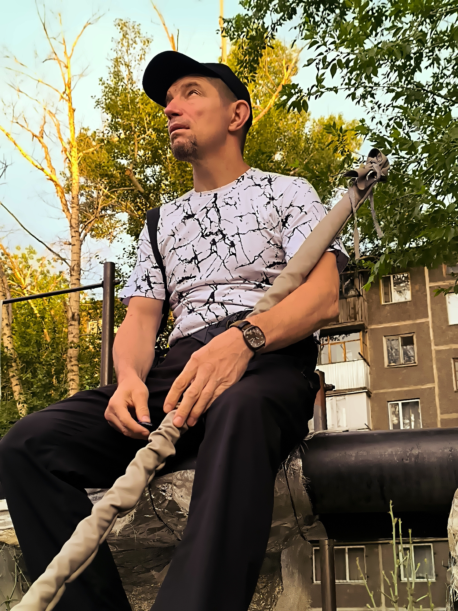 A man wearing a black baseball cap, a white t-shirt with a black abstract pattern, and black pants is sitting outdoors. He is looking upwards and to the right. He has a thin goatee. A watch is visible on his left wrist. He is holding a gray, flexible tube-like object with both hands. He is seated on a large, dark, horizontal pipe. Behind him, there is a metal railing, trees with green leaves and bare branches, and a multi-story building with balconies and windows. The setting appears to be an urban environment in the city of Qarağandı city administration, Kazakhstan. The lighting suggests it is late afternoon or early evening.