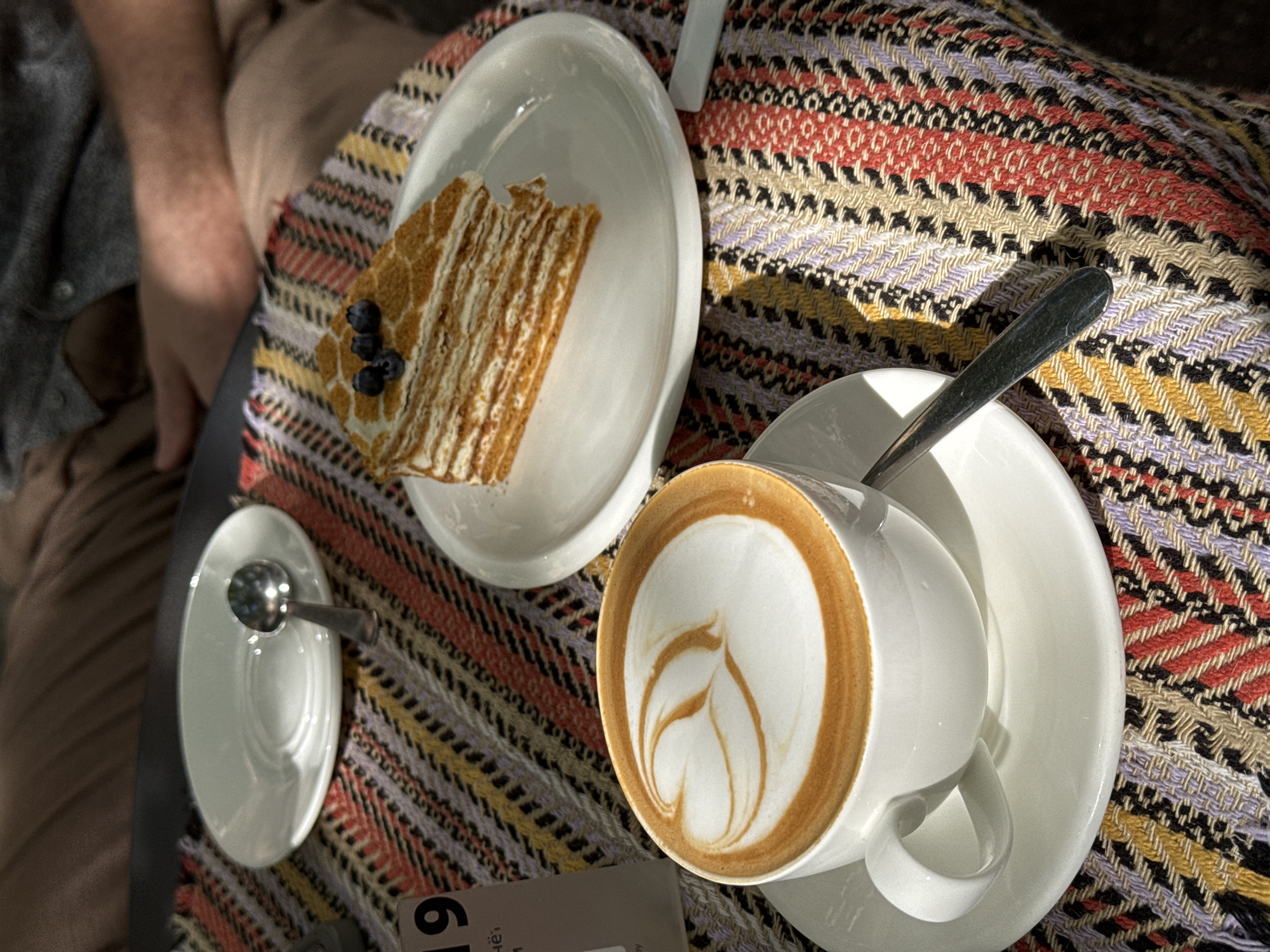 An outdoor cafe scene features a white cup of coffee with latte art on a saucer, a slice of layered cake with blueberries on a plate, and a small spoon on a patterned tablecloth. A white saucer with a spoon is to the left of the coffee. The cake is to the right of the coffee. A table number "19" is visible on the left side of the frame. The background shows a person's legs wearing tan pants and a gray shirt, suggesting they are seated at the table. The lighting indicates it is daytime with dappled sunlight. The setting is an outdoor cafe in an unknown city.