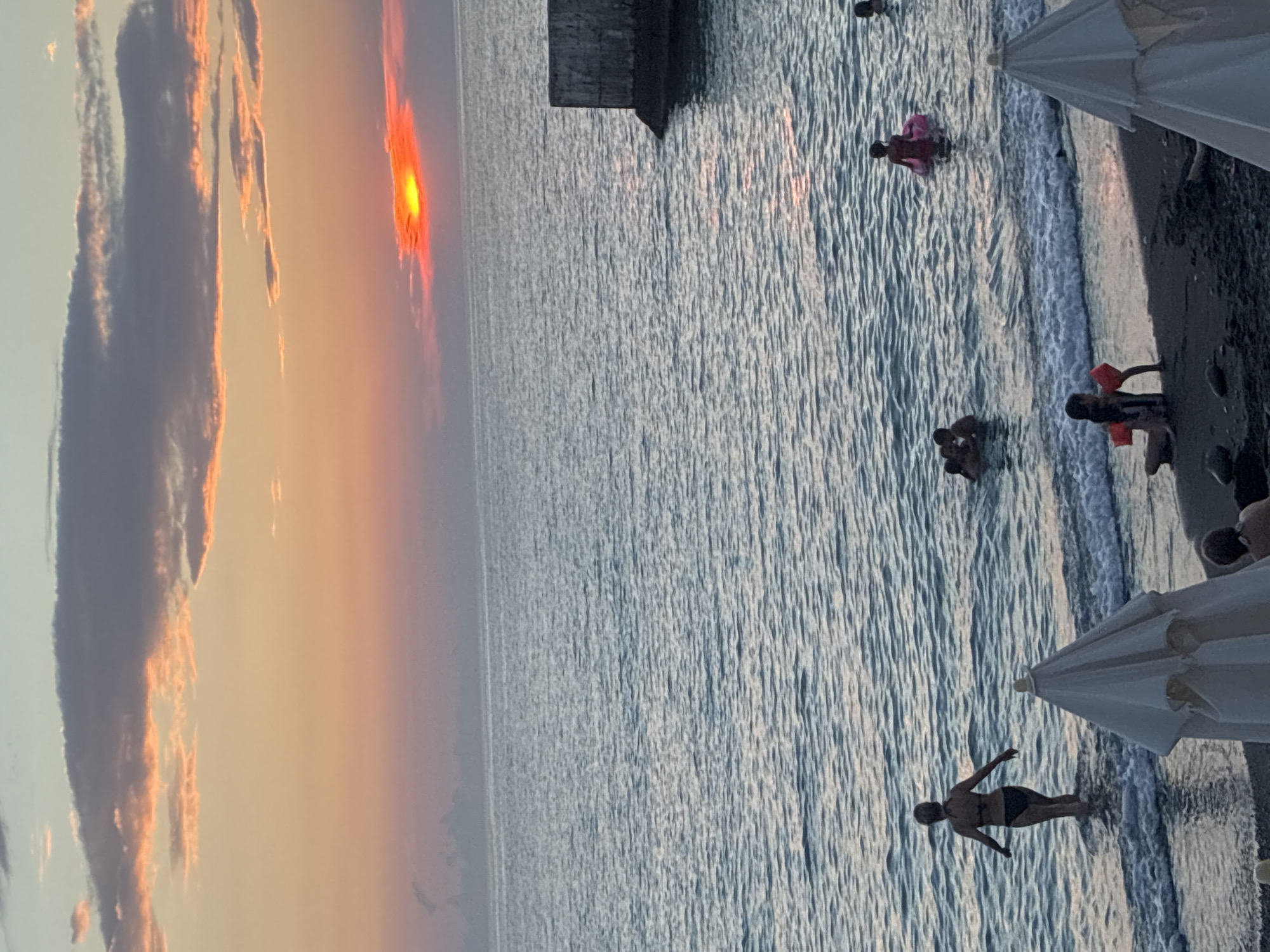 The image depicts a beach scene in Sochi, Russia, during sunset. The sky is painted with hues of orange, pink, and blue, with scattered clouds. The sun is visible as a bright orange orb, partially obscured by clouds on the horizon over the Black Sea. Gentle waves lap the shore, where several people are present.
In the foreground, a white beach umbrella is partially visible on the dark sand. Several individuals are in the water. To the left, a woman in a black bikini wades into the sea. Further out, a group of people, including children, are swimming and playing. One child is on a pink inflatable ring. On the shore, a person is sitting with orange floatation devices.
To the right of the frame, a concrete structure, possibly part of a pier or breakwater, juts into the sea. The overall atmosphere is serene, capturing a moment of leisure at the end of the day.