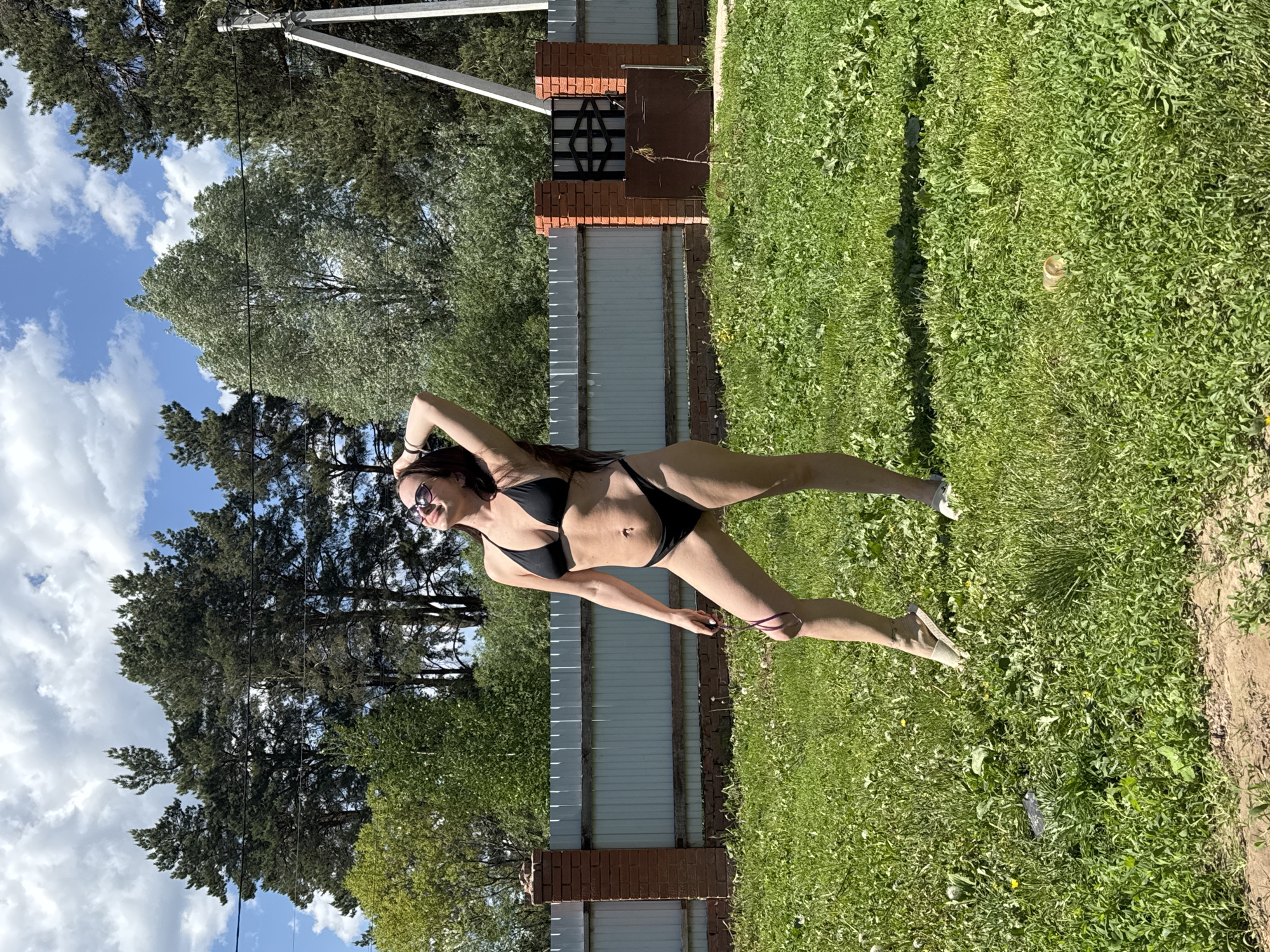A woman in a black bikini and sunglasses is posing in an outdoor setting that appears to be a backyard or garden, likely in Rusalevka, Russia. She is standing on green grass near a white fence with brick columns and a possible gate. Trees and blue sky are visible in the background. A utility pole is also visible.
