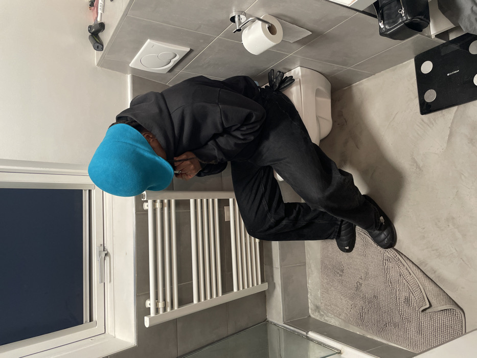 A person in a bathroom, angled and near a toilet, wears a blue hat, black hoodie, and black jeans. Their upper body is bent forward, possibly looking at something on their phone. The bathroom contains a window, a radiator, a toilet with a paper roll, a black purse, and a black scale. A bath mat lies on the floor. The scene is a slightly tilted and cropped shot.