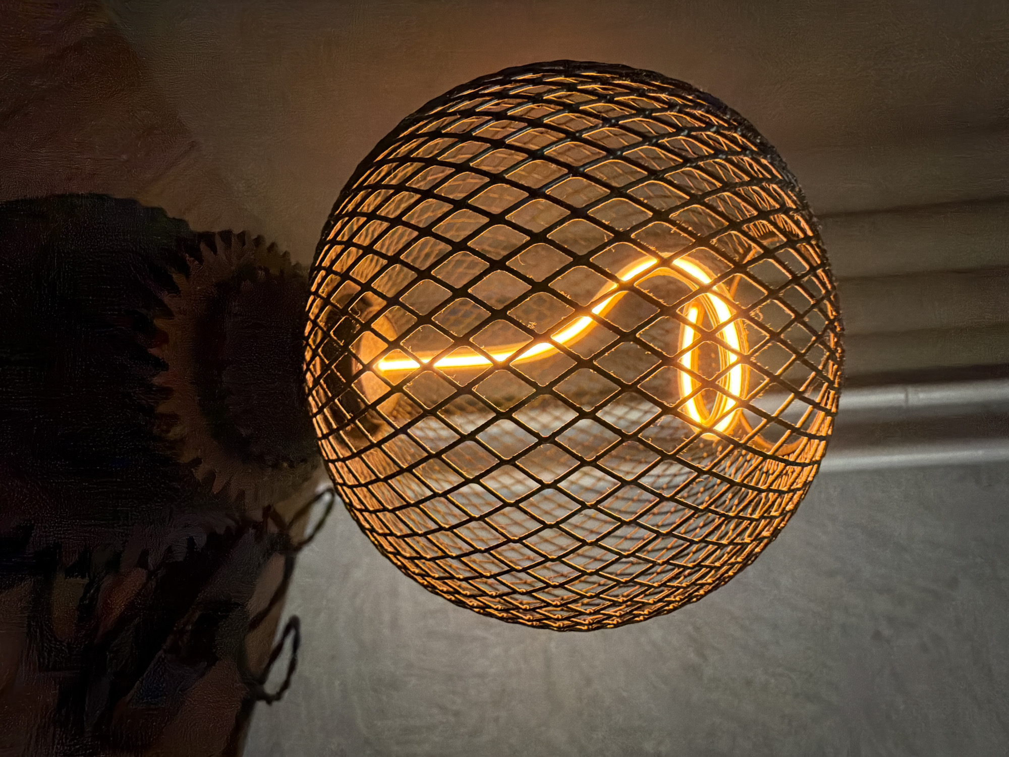 An indoor close-up shot features a spherical, industrial-style light fixture hanging prominently in the center. The fixture is made of a dark metal mesh, forming a diamond-patterned cage. Inside, a glowing filament bulb emits a warm, orange-yellow light, illuminating the intricate curves of its internal filament. The bright light causes some glare and reflections on the mesh.
The background primarily consists of textured, dark surfaces, likely a wall or ceiling. To the upper left, a shadowed, dark object with distinct gear-like or cogwheel shapes is partially visible, suggesting a decorative element or part of a larger structure. The textures in the background and on the dark object are coarse, enhancing the industrial or rustic aesthetic hinted at by the light fixture. The overall lighting is dim, indicating an evening setting or an interior space where this light source is active. No visible text is present in the image. The scene is located in Sofia, Bulgaria.
