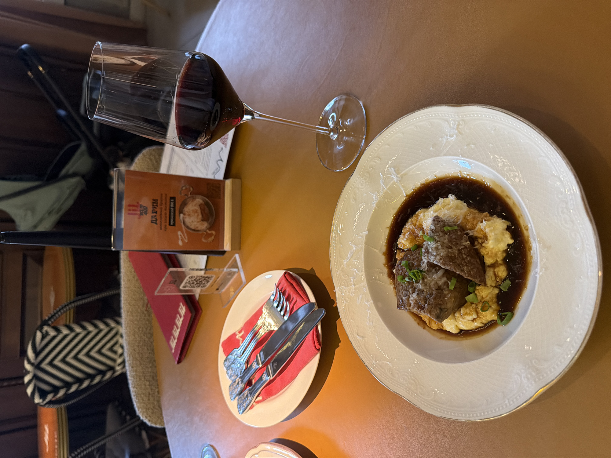 The image displays a close-up, high-angle view of a dining table setting in what appears to be a restaurant in Krasnoyarsk, Russia. The central focus is a plate of food and a glass of red wine.
The main dish consists of large pieces of braised meat, possibly beef, resting on a bed of mashed potatoes or a creamy purée, all submerged in a rich, dark brown gravy. The dish is garnished with chopped green onions and a sprinkle of grated cheese. The white ceramic plate features an elegant, embossed rim with a delicate gold accent. To the left, a stemmed glass holds a generous pour of red wine.
Adjacent to the wine glass is a brown menu book with visible Cyrillic text, reading "ДАЙ РУБИН" (Dai Rubin), likely the establishment's name. Below this, smaller text includes "МЯСО БЕЗ ИСКУССТВЕННЫХ ДОБАВОК" (Meat without artificial additives) and "ПИВОВАРЕННАЯ ФИРМА" (Brewing company). Further left, there's a small side plate with a red folded napkin holding a fork and two knives, all with ornate handles. A small acrylic stand with a QR code and a red folder, possibly also branded "RUBIN," are also present.
The table has a warm, caramel-brown surface. In the blurred background, hints of a dark chair and a patterned bag (black and white chevron) are visible, suggesting other elements of the restaurant's interior. The overall scene depicts a prepared meal, possibly for a single diner, highlighting a focus on hearty cuisine and a relaxed dining experience. The lighting is soft and warm, characteristic of an indoor setting. No people are visible in the frame.