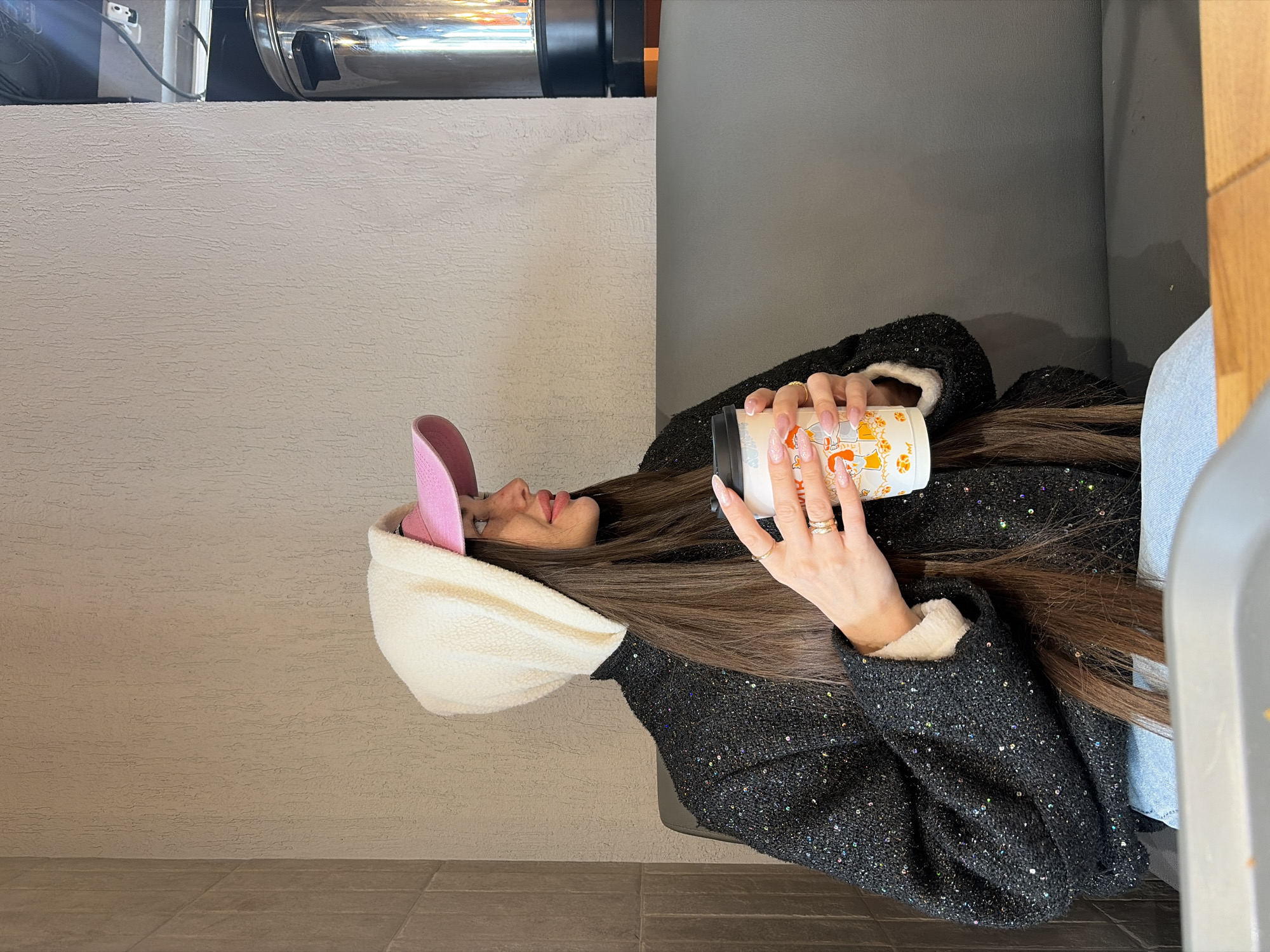 A woman with long brown hair and a pink and white hat is holding a coffee cup. She is wearing a black sparkly jacket and sitting in front of a gray wall.