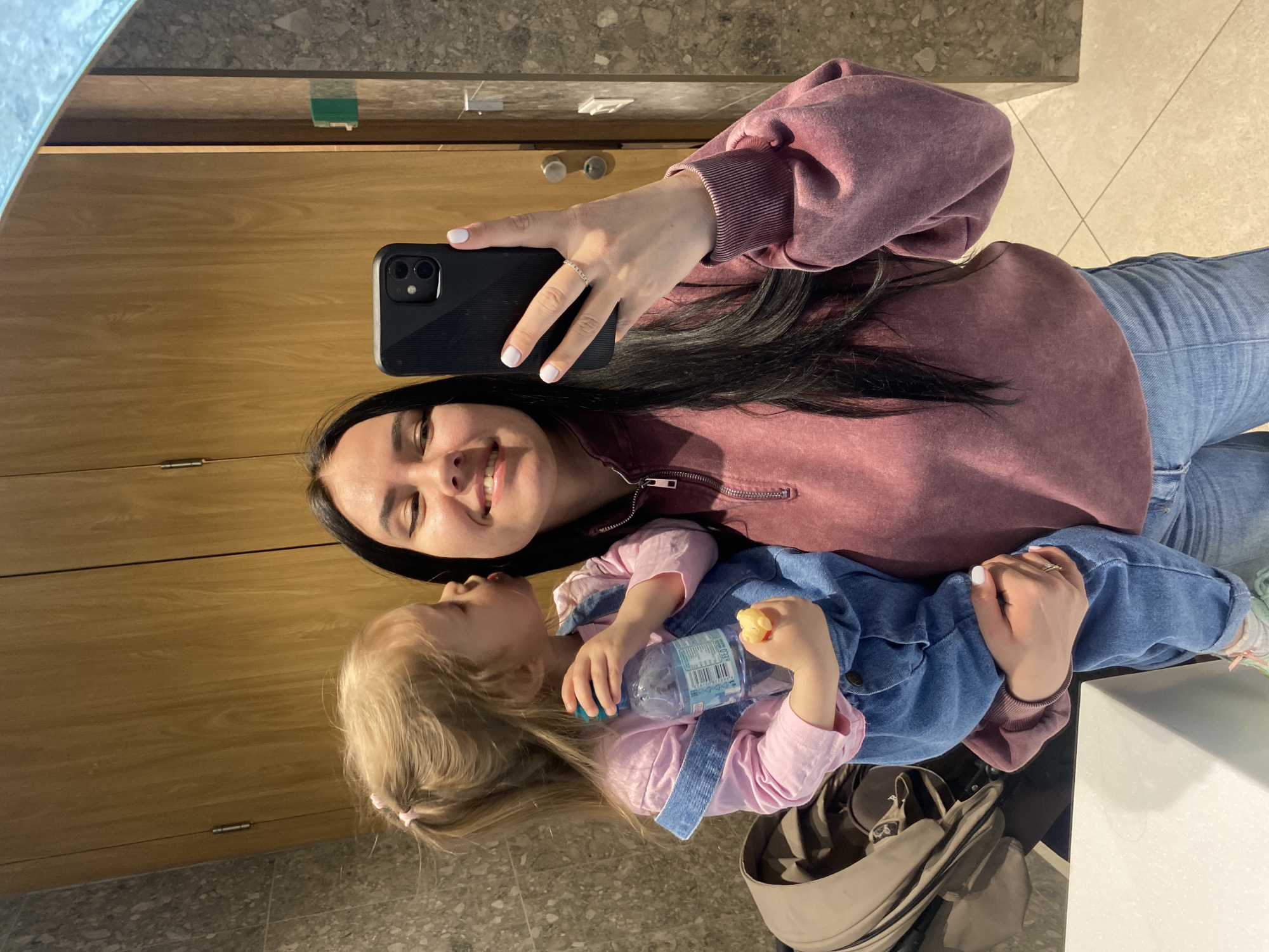 A woman and a toddler are visible in a selfie taken in a mirror. The woman has long, dark hair and is wearing a pink sweatshirt and blue jeans. She is holding a black smartphone. The toddler, who appears to be holding a water bottle and a piece of food, has blonde hair with a pink hair clip. A light brown bag is situated below them. The reflection shows wooden cabinets above a beige tiled floor. Based on visual clues, the location is in Perm, Russia.