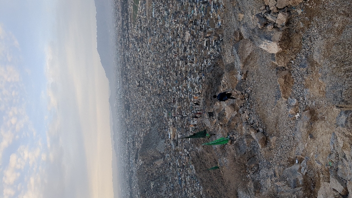 A cityscape stretches into the hazy distance below a rocky, uneven terrain. Flags and garments flutter on the hillside near a figure standing on the slope. The sky fades from a bright, cloudy horizon to a muted, dusky atmosphere above the urban sprawl. The perspective is an aerial, elevated view capturing both the natural and built environments.