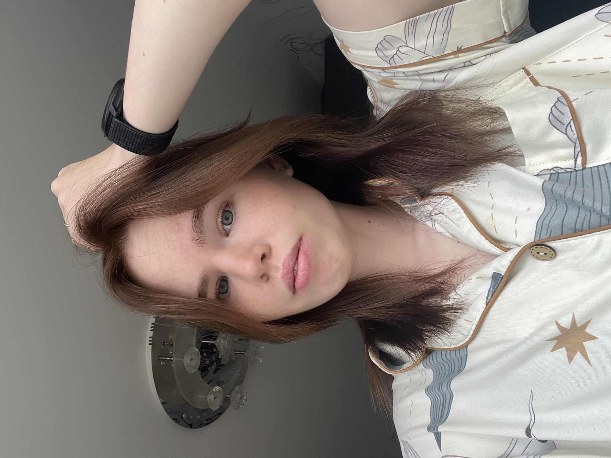 A close-up shot of a young woman with light skin, light green eyes, and light brown hair. She is wearing a pajama top that is patterned with images of birds and stars. The top has a tan trim. She has a black sports watch on her left wrist. The background is a light gray wall with a circular wall decoration, likely a light fixture. The location cannot be definitively determined as Chaikovsky, Russia.