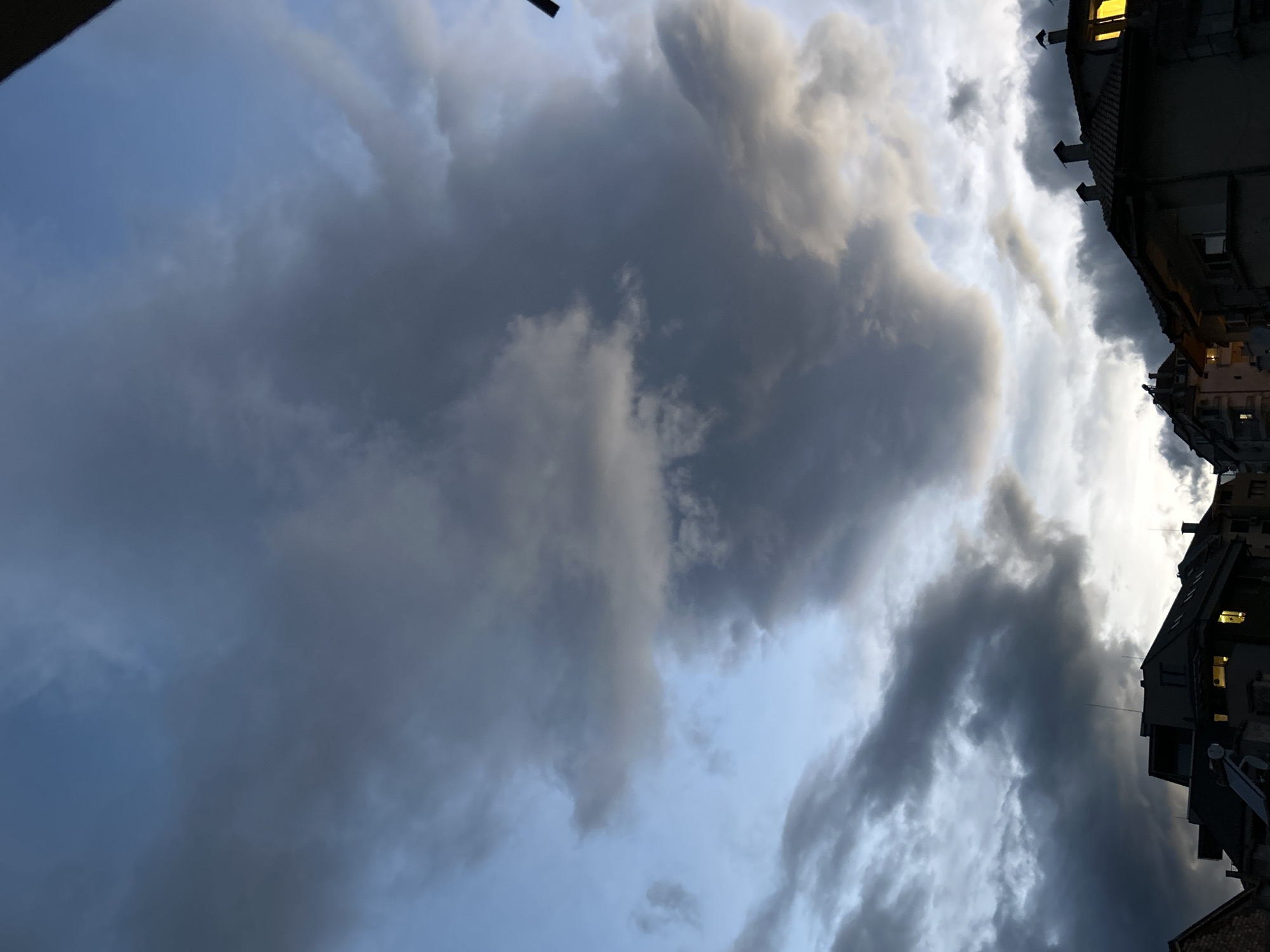 The image depicts a view of a cloudy sky with a building on the right side. The clouds are a mix of gray and white, indicating varying densities. The building is a dark silhouette, with a few windows illuminated from within. The location is in the city of Unknown, Unknown.
