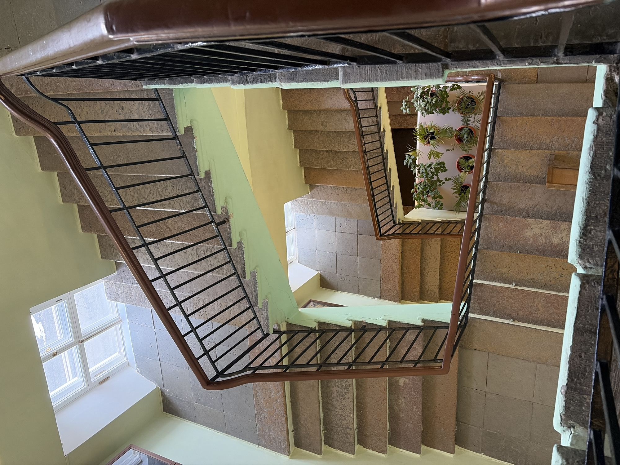 The image shows a view looking down into a stairwell of a building in Saint Petersburg, Russia. The stairwell has multiple flights of stairs with metal railings and wooden handrails. The stairs are constructed of stone or concrete. The walls of the stairwell are painted pale green and yellow. There is a window near the bottom of the stairwell, providing natural light. A vertical garden with potted plants is visible in one of the landings. The overall condition of the stairwell appears to be well-maintained with some signs of wear and tear.