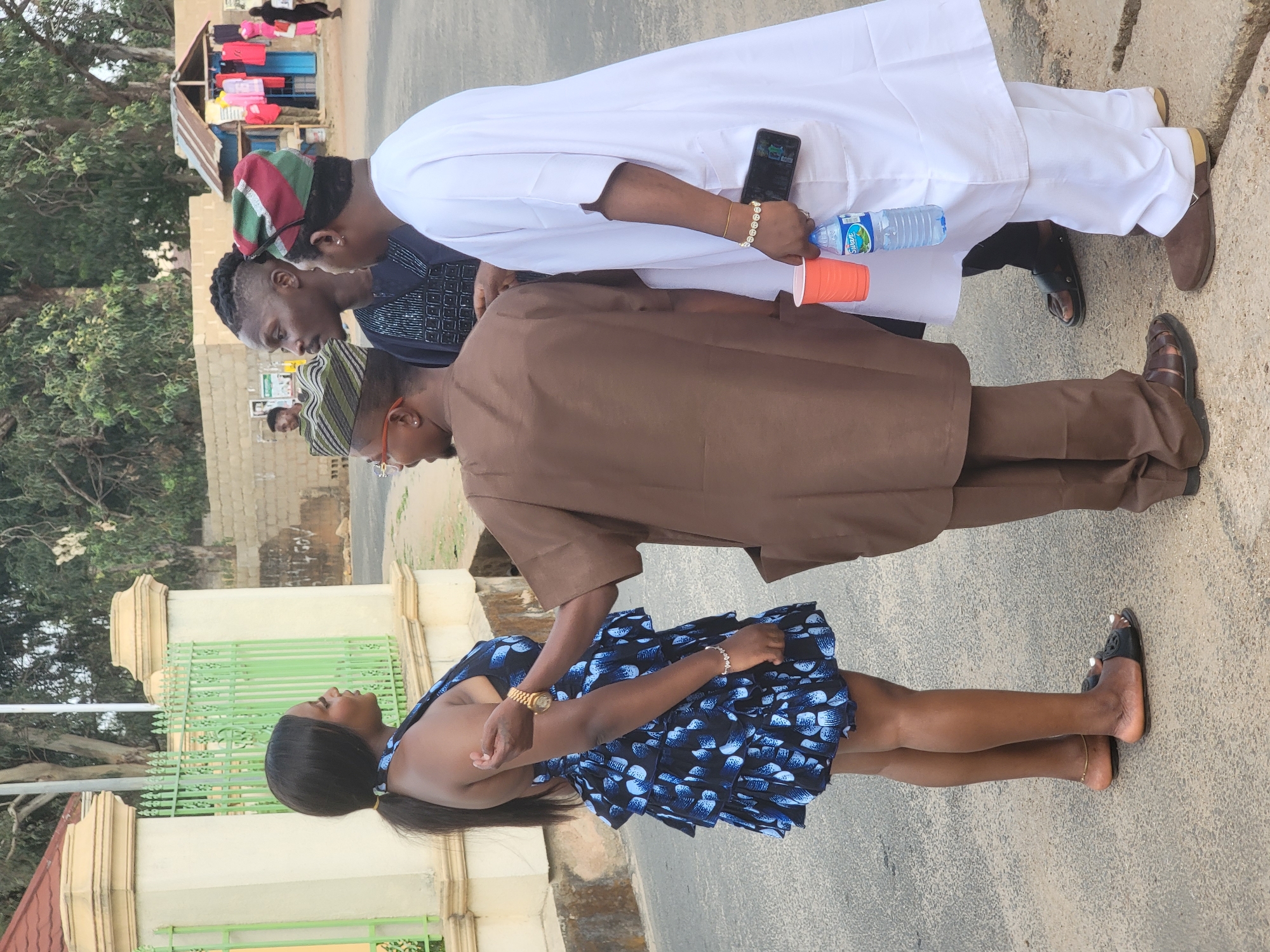 The image depicts a group of four people standing on a street in Jos, Nigeria. The background features a green and white fence, trees, and a building with a makeshift clothing rack.
A woman in a short blue and black patterned dress stands in the foreground. A man wearing a brown traditional outfit is standing next to her. Two men, one in a white traditional outfit holding a water bottle and a phone, and another in a dark patterned shirt and a cap, stand adjacent to each other. The group seems to be engaged in a conversation or observation. The ground is paved.