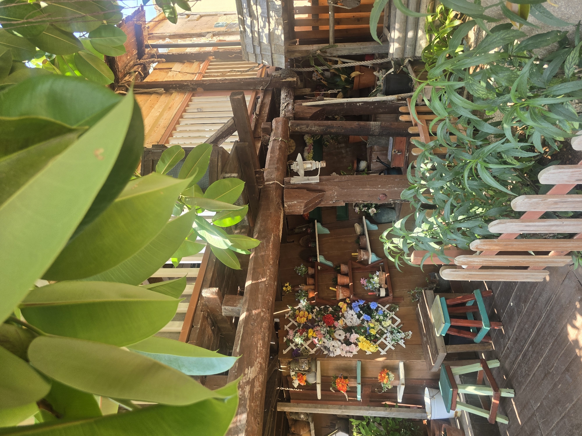 This image depicts a rustic, multi-level wooden structure resembling a treehouse or a stilted dwelling, set in a lush, green environment. The structure is adorned with numerous potted plants and artificial flowers, creating a vibrant, garden-like atmosphere. Several small, brightly painted wooden stools are placed on the wooden deck in the foreground. A small, decorative wooden fence separates the deck area from a bed of dense green foliage. The overall impression is one of a charming, bohemian, and slightly overgrown outdoor living space. While the image is detailed, it does not provide any specific visual cues to pinpoint its exact location within the city of Mellieħa, Malta.
