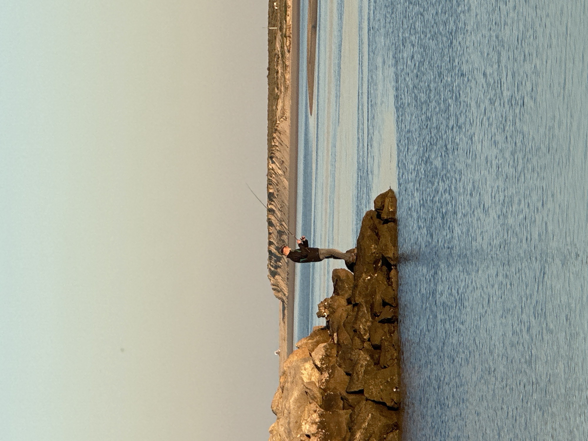 A man is fishing from a rocky outcrop on the coast of Olhão, Portugal. The man is wearing a dark jacket and trousers and a hat. He is holding a fishing rod and looking out to sea. The rocky outcrop is surrounded by water. The background is a coastline of sandy beaches and dunes. The sky is overcast.