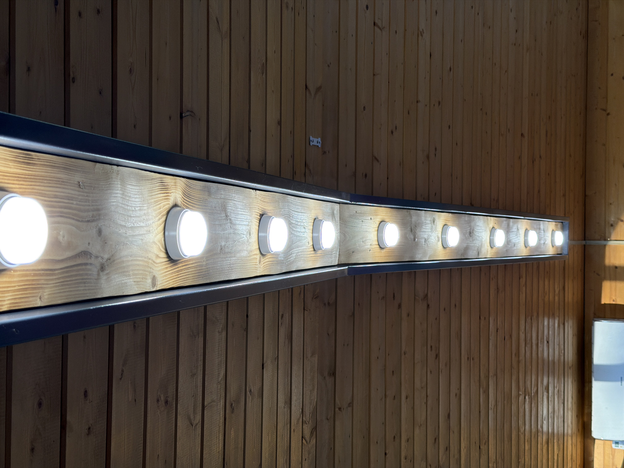 The image shows a long, linear light fixture mounted on a wooden wall. The fixture is rectangular with a dark-colored metal frame and a wooden interior. It contains a series of round, white light bulbs, evenly spaced along its length. The bulbs are illuminated, casting a bright, white light.
The wall behind the fixture is constructed from vertical wooden planks. The wood has a natural, light brown color with visible grain and knots. The lighting suggests an indoor setting, possibly a hallway, studio, or a functional space like a backstage area. The overall aesthetic is simple and functional, with the warm tones of the wood contrasting with the bright, modern lights.
There is a white electrical box visible on the far right side of the image, mounted on the wooden wall. A small white bracket with two holes is also visible on the wooden wall in the upper middle portion of the image.
The scene is static, with no people or activity depicted. The focus is solely on the architectural elements and the lighting fixture. The time of day cannot be determined from the image. The location context provided is Bol'shaya Ven'ya, Russia. There is no visible text in the image, other than potentially small labels on the electrical box, which are not legible.