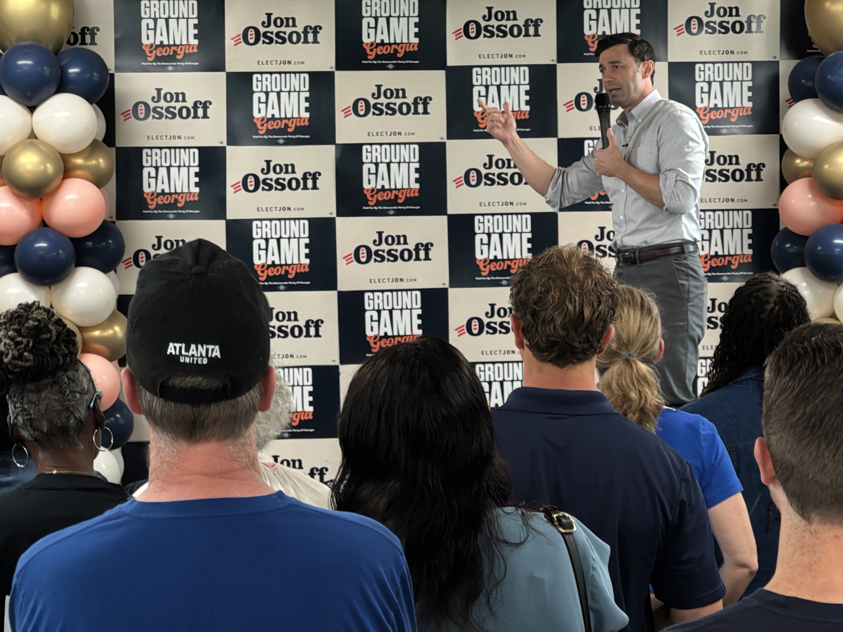 Here's a detailed description of the image:
**Scene Overview:**
The image captures a campaign event for Jon Ossoff, likely taking place indoors. A crowd of people is gathered, listening to Ossoff, who is standing and speaking into a microphone.
**People:**
* **Jon Ossoff:** He is the main focus, standing in the upper right. He is speaking and gesturing.
* **Audience:** A group of people, primarily viewed from the back, is listening to Ossoff.
**Objects:**
* **Balloons:** There is a cluster of balloons on the left side of the frame in colors of gold, white, pink, and navy blue.
* **Campaign Backdrop:** A backdrop with the "Jon Ossoff" name, "Ground Game Georgia", and his website "ELECTJON.COM" is visible behind him, in a repeating pattern.
* **Microphone:** Jon Ossoff is holding a microphone.
* **Hat:** A person in the audience is wearing a black hat with "ATLANTA UNITED" printed on the back.
**Actions/Interactions:**
* **Speaking:** Jon Ossoff is giving a speech or presentation.
* **Listening:** The audience is engaged in listening to the speaker.
**Environment:**
* The environment appears to be indoors, possibly a meeting room or event space.
* Based on the details provided in the OCR output, it seems to be an event in Decatur, Georgia, USA.