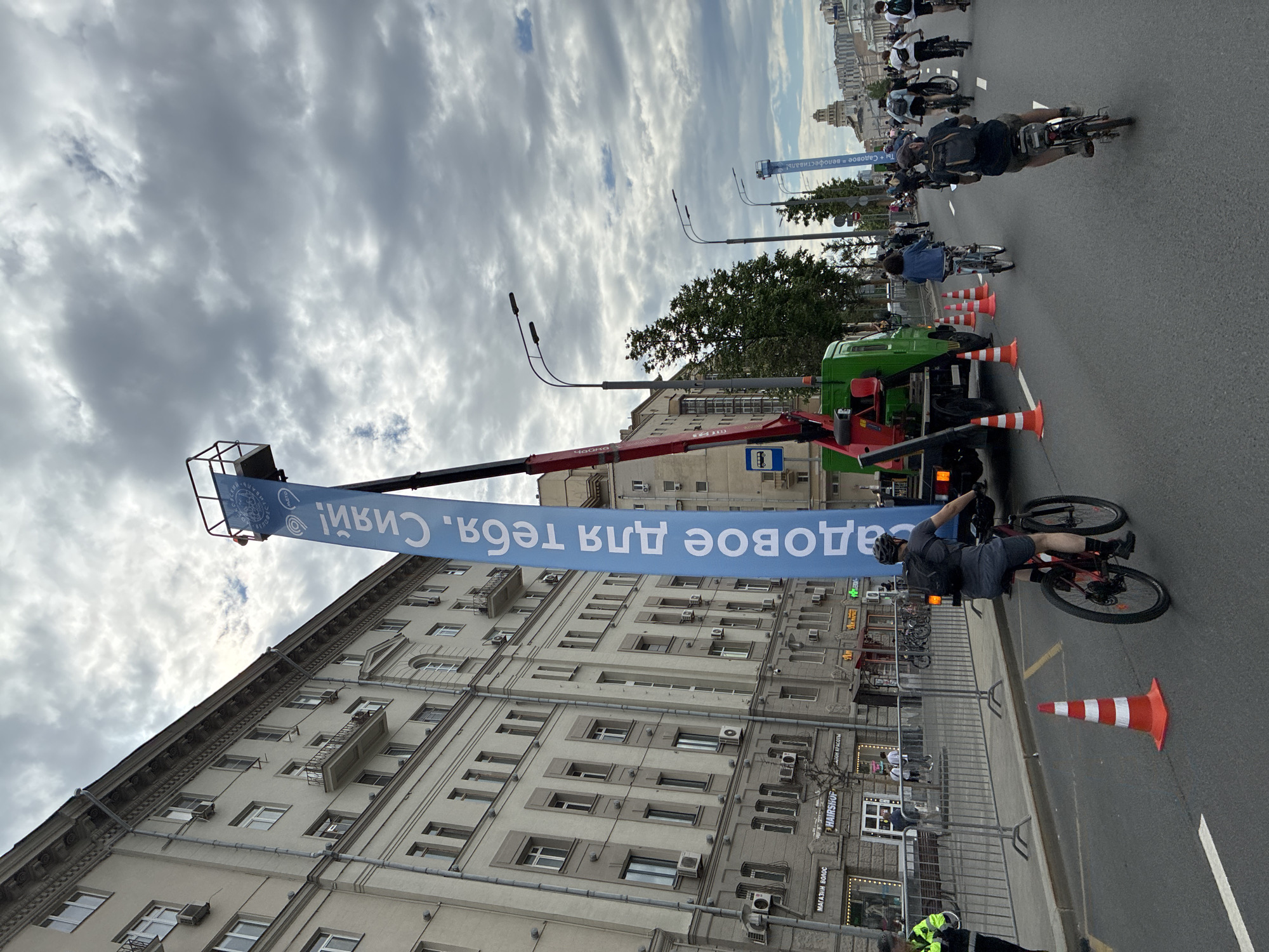 The image depicts a scene on a city street, likely in Moscow, Russia, suggested by the style of buildings and the Cyrillic script on the banner and storefronts. A large banner, mounted on a mechanical lift extending from a green truck, stretches across the road. The banner reads, "Садовое для тебя! Сияй!" (Sadovoe for you! Shine!).
The street is filled with cyclists participating in what appears to be a cycling event. Orange cones are placed on the road, potentially marking the route. A cyclist is passing by the truck.
The buildings on the side of the street are tall, multi-story structures with numerous windows and architectural details. Several storefronts are visible at street level. The sky is overcast with cloudy conditions.