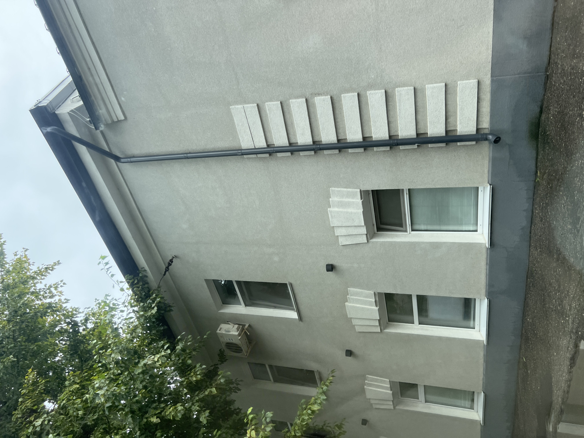 This image depicts the side of a light gray, multi-story building located in Ulyanovsk, Russia. The building has several windows with white frames. Some windows have blinds. An air conditioning unit is mounted on the wall to the left of the largest window. A dark gray downspout runs vertically along the side of the building. To the right of the downspout, there are decorative horizontal rectangular elements on the wall, arranged in a staggered pattern. A portion of a leafy tree is visible in the upper left corner of the image, obscuring some of the building's upper section. The ground in the foreground appears to be asphalt and is wet, suggesting recent rain, with reflections visible on the surface. The sky is overcast.