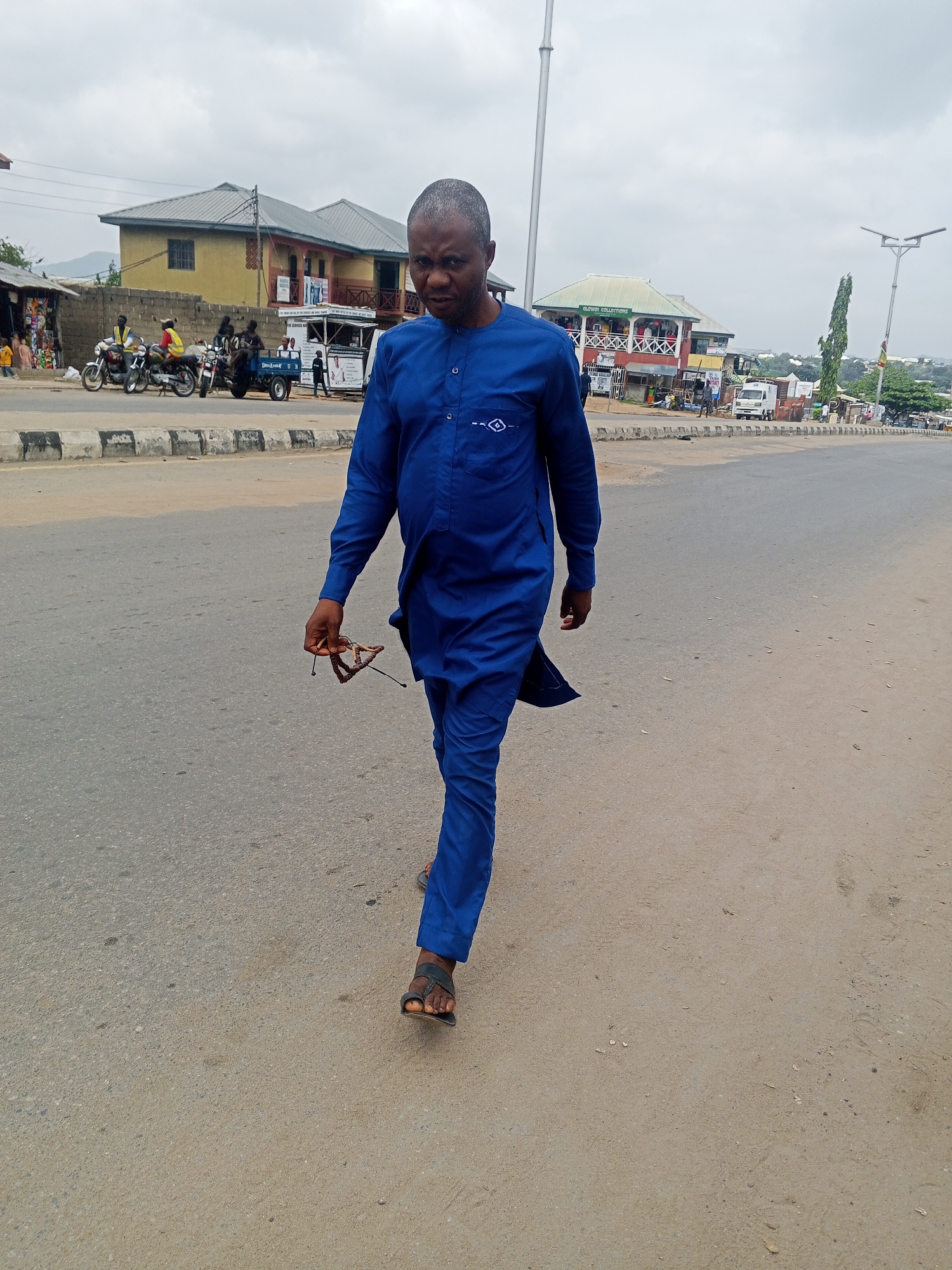 In Zango-ndaji, Nigeria, a man walks down a street. He is wearing a blue traditional outfit and sandals. In his right hand, he holds a pair of glasses. The street is paved and appears to be a busy area, with shops and motorbikes visible in the background. The sky is overcast, suggesting a cloudy day.