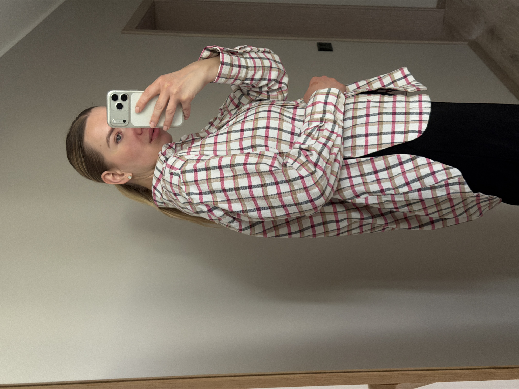 A woman is taking a selfie in a mirror. The woman is wearing a pink and brown plaid shirt and a black skirt. She is holding a white smartphone. The background includes a wall and a doorway. There is no evidence to establish that this scene is located in the city of Dreiliņi, Latvia.