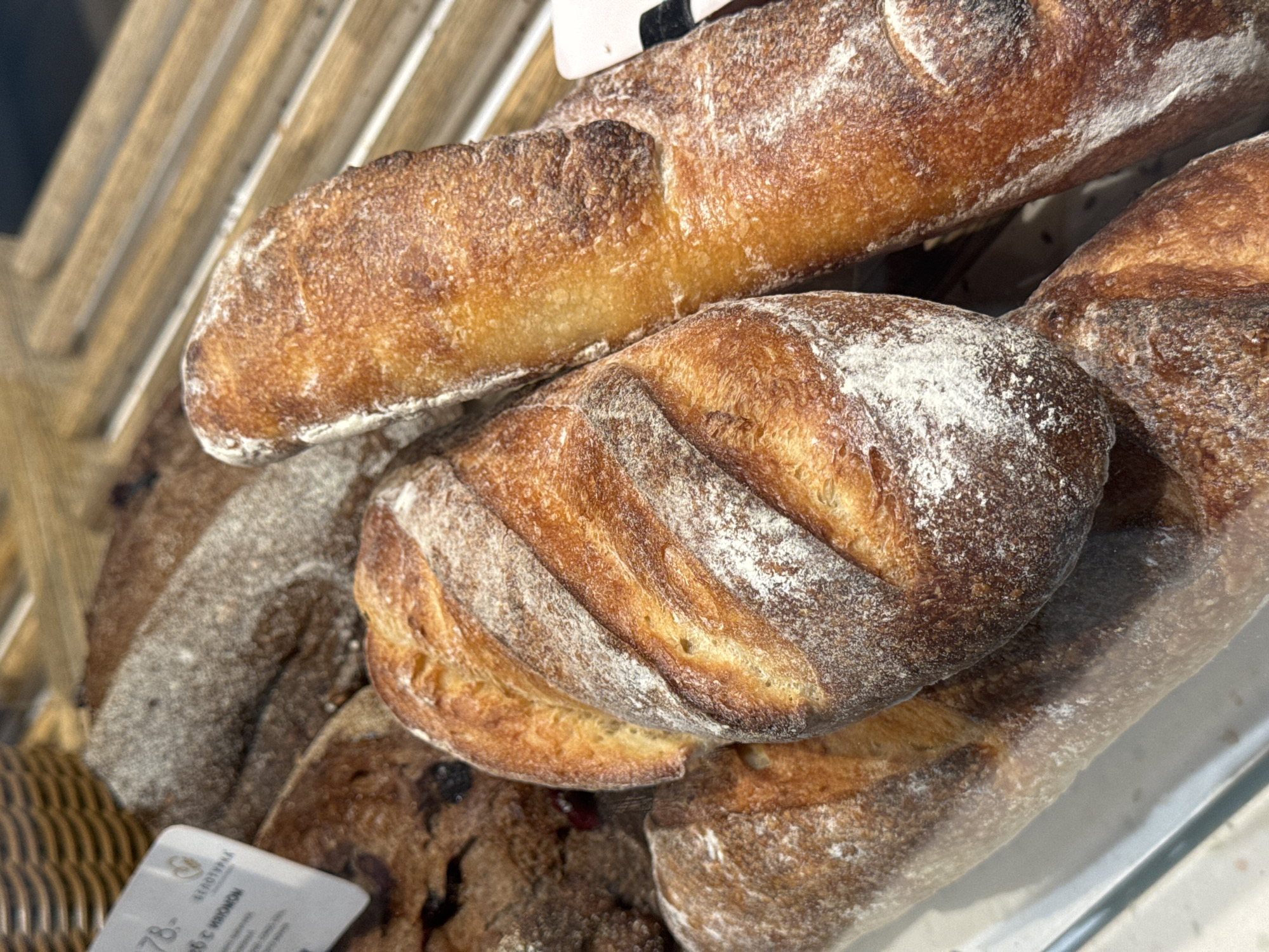 A close-up shot shows an assortment of breads in a display case, dusted with flour. The breads are of various shapes and sizes, including a long baguette-style loaf and a round loaf with scoring. The lower left corner shows a corner of a wicker basket, with a price tag in Russian visible, suggesting a location in a bakery or grocery store in Izhevsk, Russia. The overall impression is of fresh, artisan breads.