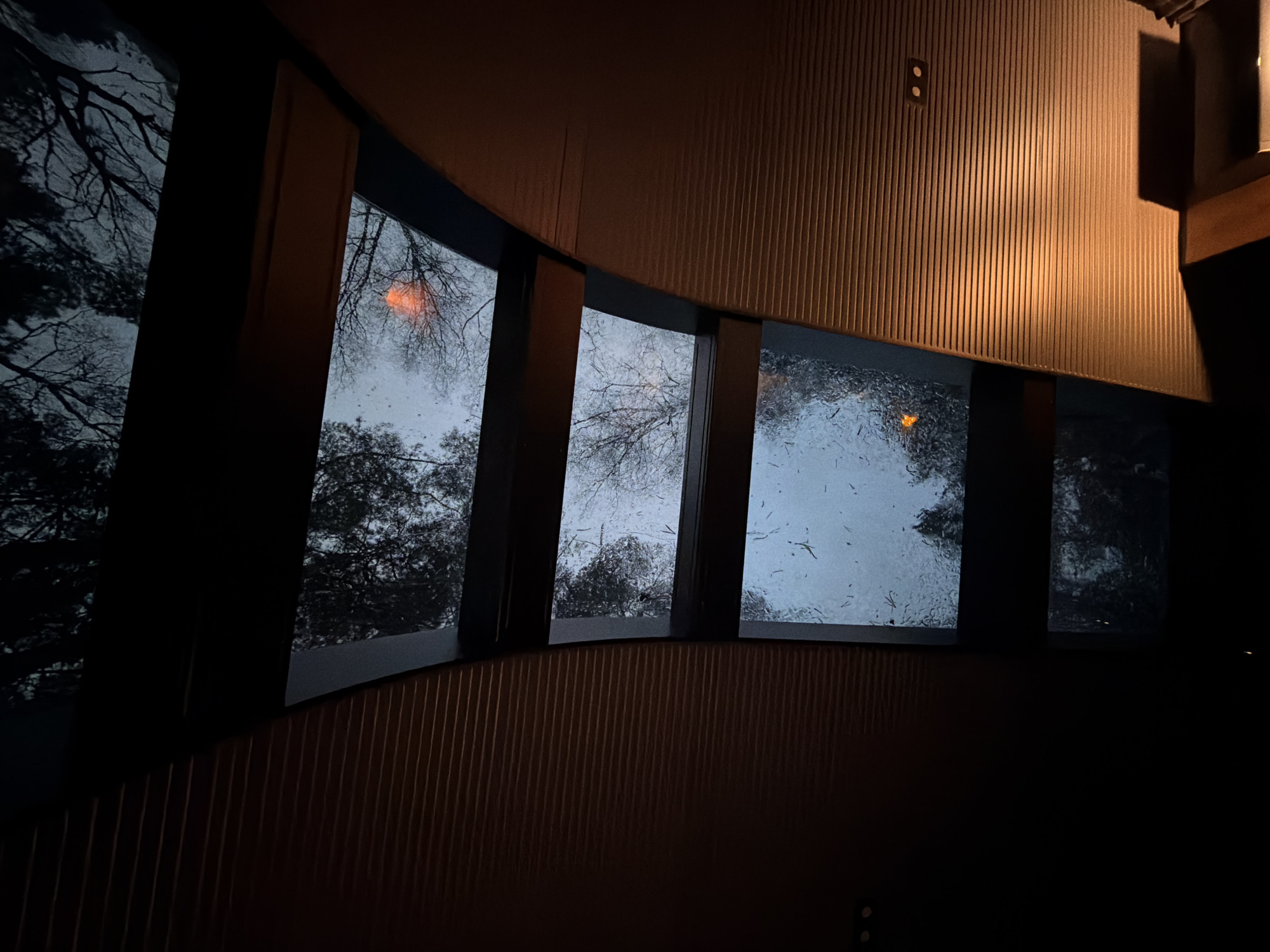 The image depicts an interior view looking out through a series of windows. Outside, the sky is overcast with trees visible, some appearing bare. Rain or moisture is present on the windows. The interior features a wall with a grooved, possibly wooden, texture. An electrical outlet is visible on the wall. The scene is dimly lit. The location in the city of Санатория "Восход", Russia is not possible to verify with the given details.