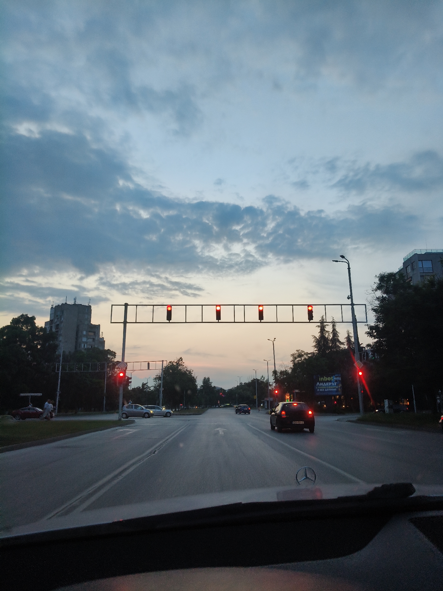 The image captures an urban street scene at dusk, specifically at the intersection of Bulgaria Boulevard and Vasil Aprilov Boulevard in Plovdiv, Bulgaria, viewed from the driver's perspective of a Mercedes-Benz vehicle (indicated by the hood ornament). The sky overhead is predominantly blue-grey with scattered darker clouds, transitioning to a lighter, yellowish-orange hue near the horizon, indicative of twilight.
The vehicle is stopped at a red traffic light. A prominent overhead gantry spans the width of the road, displaying four red circular traffic lights illuminating brightly. An additional red traffic light is visible on a pole to the left, and another on a pole to the far right. The asphalt road surface appears damp, reflecting the overhead lights faintly, and features a distinct double white line separating lanes, along with other faded white lane markings.
Several other vehicles are stopped ahead at the intersection. Immediately ahead and slightly to the left, a dark-colored hatchback with Bulgarian license plates "PB 4408 AC" is visible, its taillights illuminated. To its left, a silver sedan is stopped, and further left, a grey sedan. In the distance, a blue car is also visible. On the left side of the road, near a parked red car, two individuals are standing on the sidewalk or shoulder.
The environment features various buildings and vegetation. On the left, a multi-story, older concrete apartment building with several antenna structures on its roof is partially obscured by dense green trees. On the right, a more modern building with dark windows and a flat roof is visible above a cluster of trees. A prominent billboard sign is positioned on the right side of the road, displaying text in Bulgarian: "inbet ЛИДЕРЪТ с НОВИ ОФЕРТИ И БЕЗ ДЕПОЗИТ" (inbet THE LEADER with NEW OFFERS AND NO DEPOSIT). Streetlights with curved poles are spaced along the road, and additional street furniture and poles are present throughout the scene. The overall impression is one of a busy urban intersection during the early evening hours.