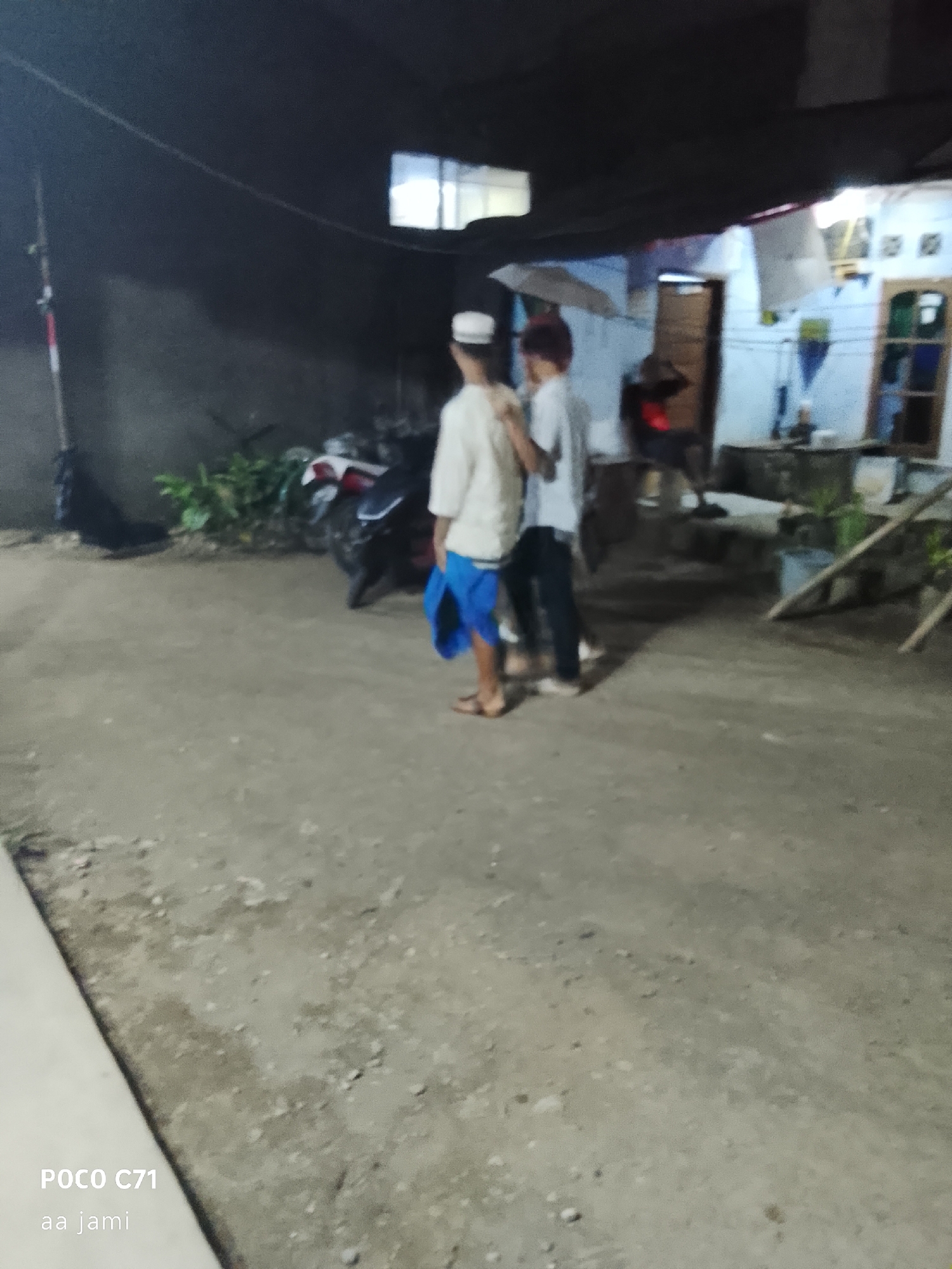 The image shows two young males in what appears to be a rural or semi-urban residential area at dusk or night. One male on the left wears a white shirt and blue shorts and a white cap. The other male on the right wears a white shirt and dark pants. They are standing in a dirt or gravel courtyard. Behind them, a motorcycle is parked near some vegetation and a wall. Further in the background, a simple house with a window is visible. The overall scene suggests a casual, everyday moment. The presence of the "POCO C71" watermark indicates the image was captured with a Poco C71 smartphone. Without additional information, it is impossible to confirm the location within Tasikmalaya, Indonesia.