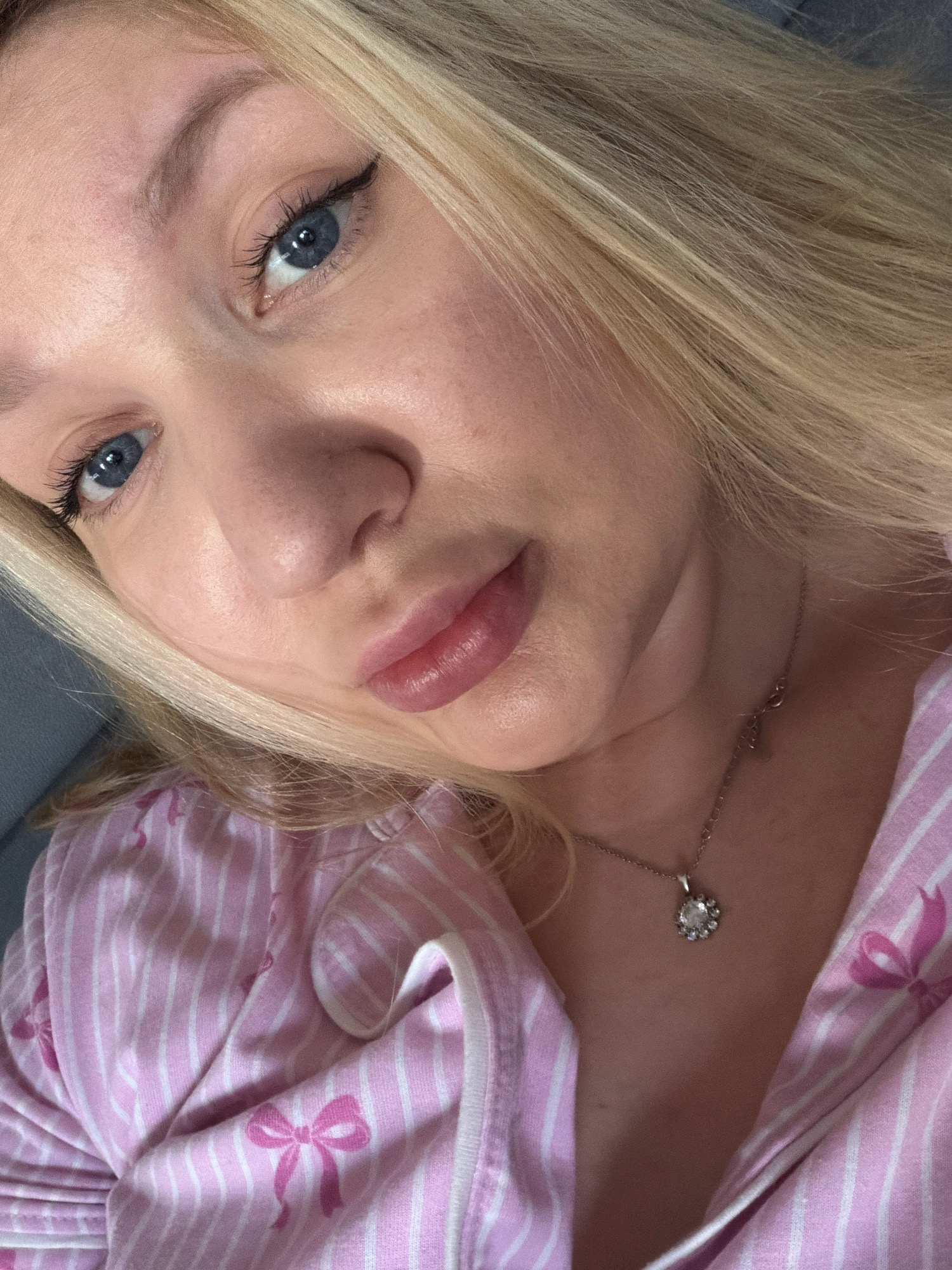 The image is a close-up, head and shoulders shot of a young Caucasian woman with blonde hair and blue eyes. She is wearing a pink and white striped pajama top with a bow print. A silver necklace with a circular pendant is visible around her neck. The lighting is soft, and the background appears to be a blurred indoor environment. The photo is taken from a slightly high angle, looking down at the woman. There are no discernible elements in the image that would allow for the determination of a specific location in the city of Koszalin, Poland.