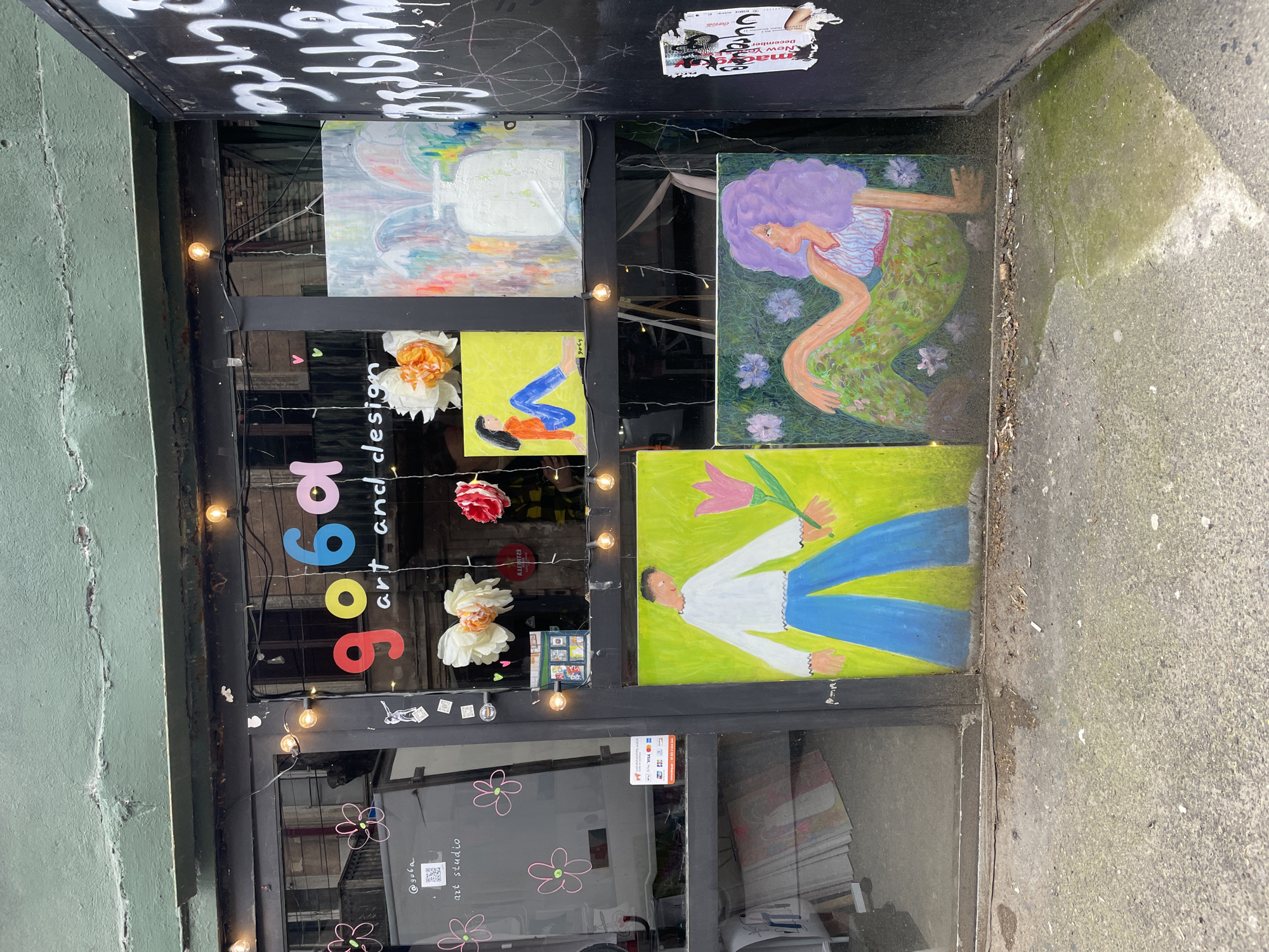 A street-level view of a shop window in Tbilisi, Georgia, displays the name "9696 art and design" vertically. The shop front is framed in dark metal. Various paintings are mounted inside the window. One shows a person in blue jeans and a white shirt holding a tulip. Another depicts a figure with lavender hair in a mermaid-like pose against a green background with flowers. Other artwork is also visible. Paper flower decorations are placed among the paintings. A string of small lights hangs across the window. Graffiti is scrawled on the wall above the shop. The sidewalk is concrete and slightly weathered.