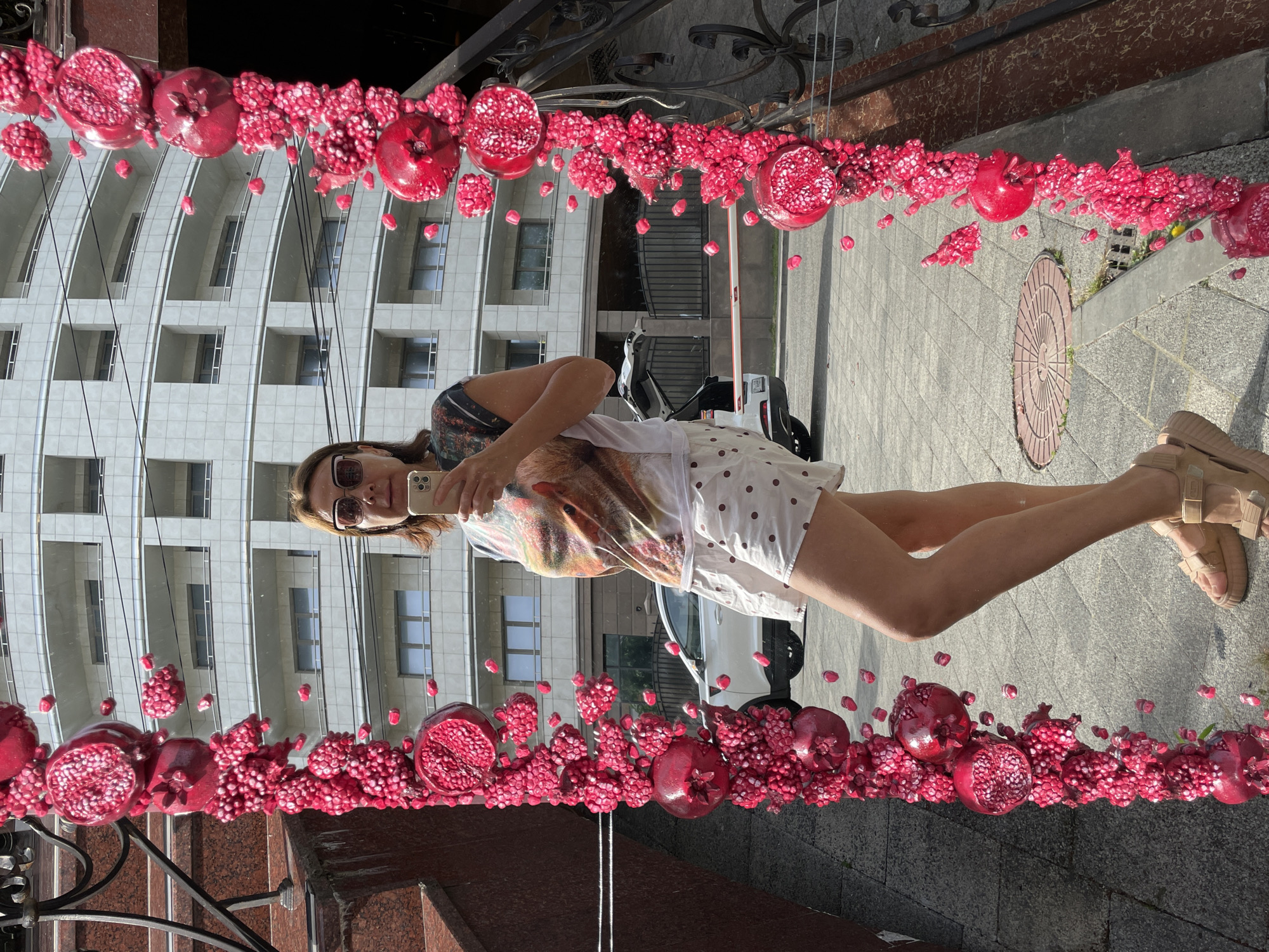 A woman, approximately in her 40s, stands in front of a building in Sochi, Russia, taking a mirror selfie. She is wearing a white t-shirt with a colorful graphic print and white shorts with polka dots, along with tan platform sandals. She is holding a smartphone in her right hand. The mirror is decorated with artificial pomegranates and pomegranate seeds, creating a border around the reflection. In the background, a modern, multi-story building with balconies and large windows is visible. A white SUV with its trunk open is parked on the paved area in front of the building. A black metal fence and stairs are also present in the foreground. The scene appears to be outdoors on a sunny day.