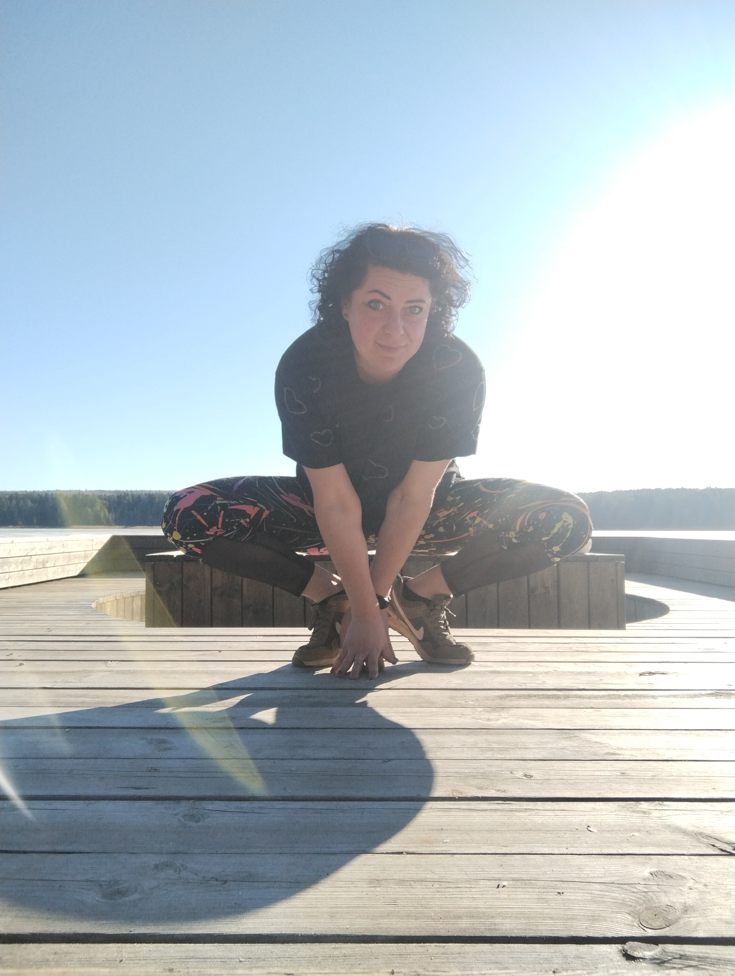 A woman with curly dark hair is squatting on a wooden deck. She wears a black t-shirt with small white heart patterns, leggings with a colorful abstract print, and tan sneakers. Her hands are resting on the deck in front of her. The background includes a lake and a line of trees. The sky is clear and blue with a bright sun in the upper right corner. The scene is brightly lit, creating strong shadows on the wooden deck.
There is no information that shows location being Votkinsk, Russia.