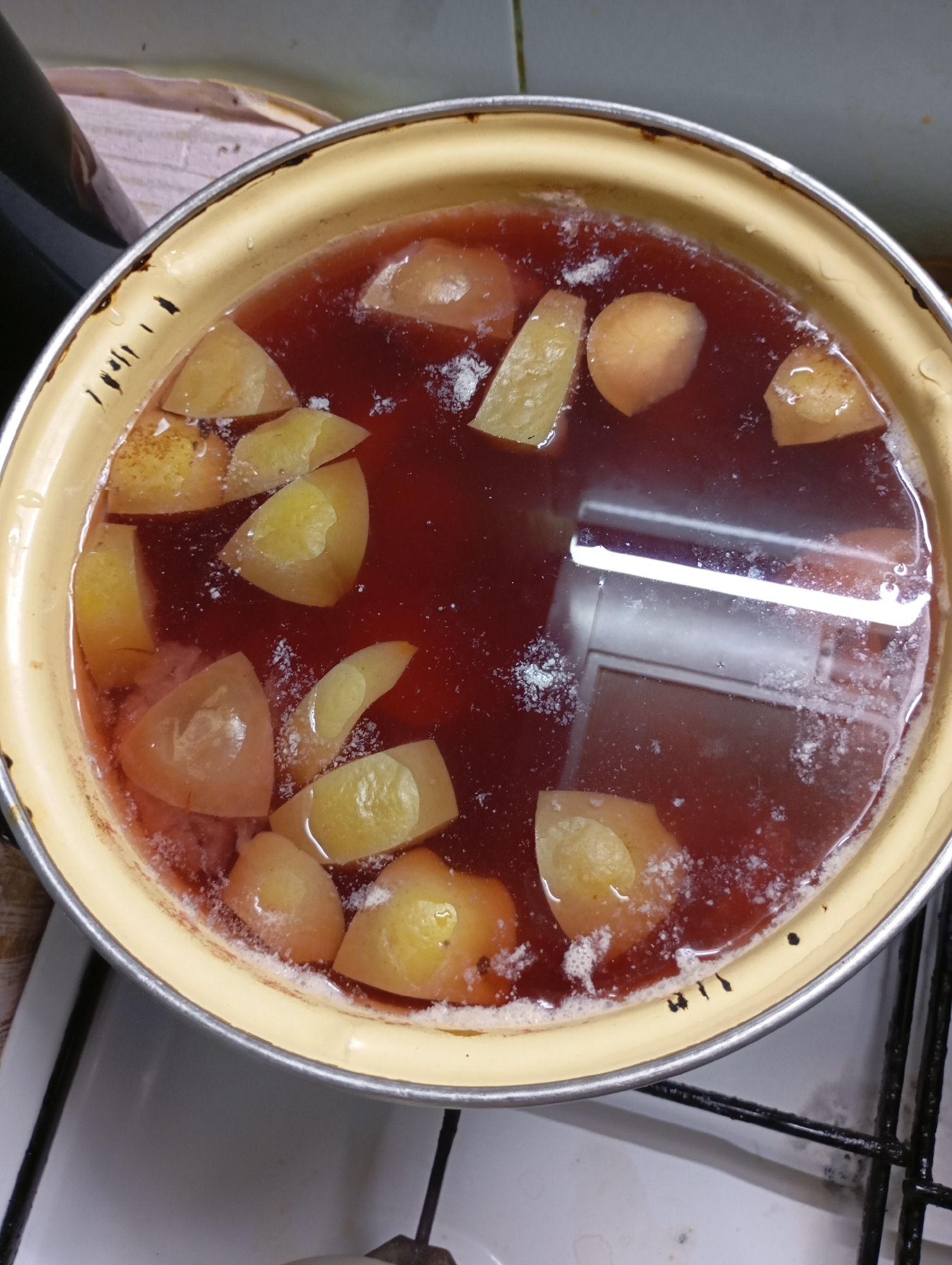 A top-down, slightly angled medium shot captures a pot of fruit simmering on a stovetop. The pot, a light yellow enamelware with a silver rim, is filled with a dark red liquid and pieces of light yellow fruit, possibly apples or pears, cut into wedges. Some white foam or residue is visible on the surface of the liquid. The pot is positioned on a black metal stovetop grate. The background shows a white tiled wall and a hint of a patterned textile to the left. The scene is domestic, likely a kitchen. No specific location in Horishni Plavni, Ukraine is discernible from the image.