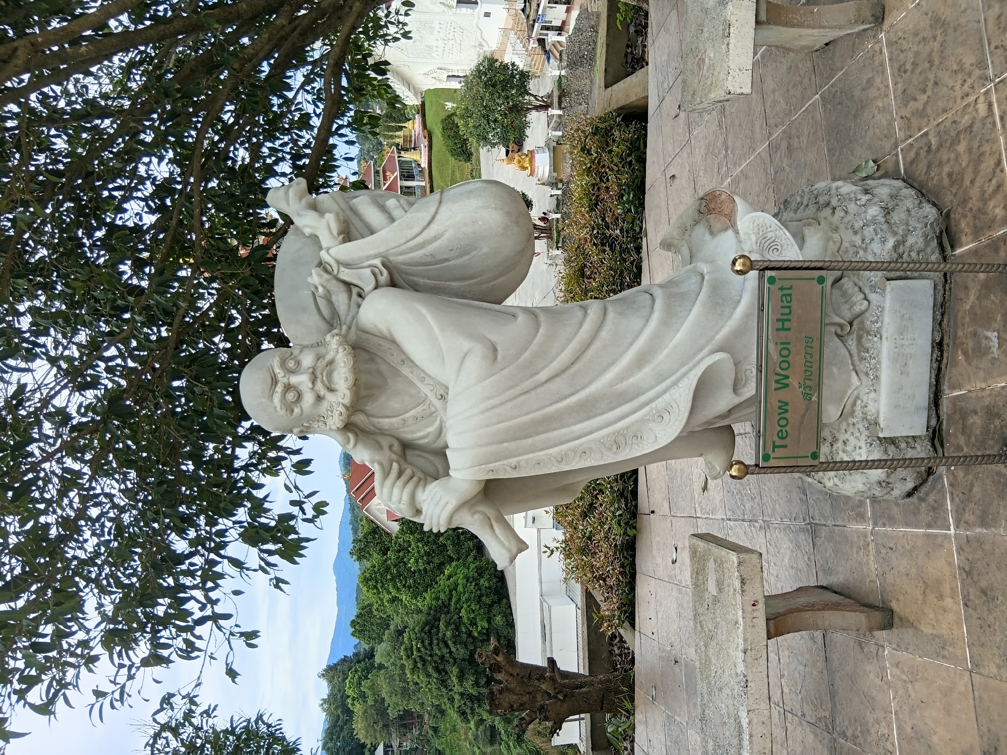 The image shows a large white marble statue of an elderly man with a long beard, wearing a robe and carrying a large sack over his shoulder. The statue is positioned outdoors on a paved area, with a sign in front of it that reads "Teow Wooi Huat". In the background, there are lush green trees, mountains, and buildings that appear to be part of a temple complex, with distinctive tiered roofs. Two stone benches are visible on either side of the statue. The sky is overcast. The overall scene suggests a serene and spiritual setting, likely a tourist or religious site in Ban Lao Phatthana, Thailand.