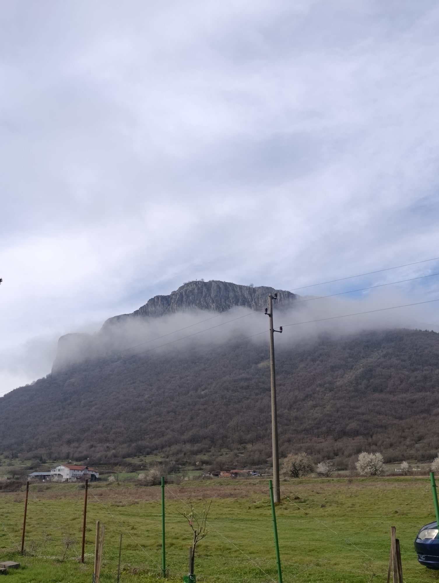 The image displays a mountainous rural landscape in Shiroko pole, Bulgaria, under an overcast sky featuring light grey clouds with intermittent patches of blue. A large, rocky mountain dominates the background, its upper half partially obscured by low-hanging mist or clouds. The mountain's lower slopes are densely covered with leafless deciduous trees, suggesting a late winter or early spring season.
In the foreground, a vibrant green grassy field is present. A fence, composed of both dark brown wooden posts and bright green metal posts strung with wire, runs horizontally across the lower frame. A tall, light-colored utility pole stands vertically in the mid-ground on the right, equipped with insulators and carrying multiple power lines that extend horizontally across the scene.
Further back, at the base of the mountain, several buildings are visible, including a multi-story white house with a prominent red roof and other smaller structures. Scattered around these buildings are several trees displaying white blossoms. The front right quarter of a dark blue passenger vehicle is partially visible in the bottom right corner of the frame. No individuals are depicted.